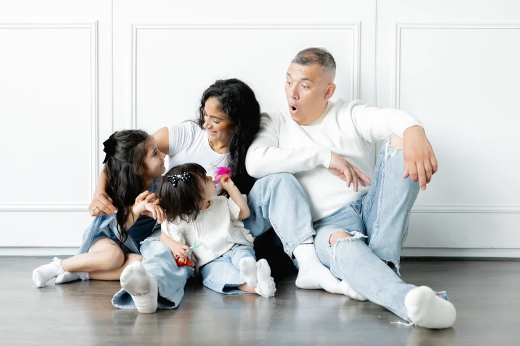 A mother, father and two little girls in white and denim sit on a studio floor, laughing, while dad pulls a surprised face.