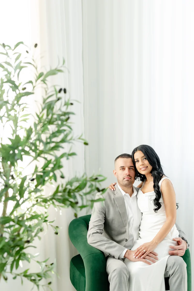 An engaged couple in soft white and grey sit close together on a green velvet chair beside an olive tree, in a bright studio with sheer curtains.