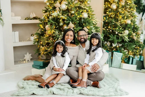 A mother, father and two young daughters in matching neutrals sit on a faux-fur rug in front of two lit Christmas trees and wrapped gifts.