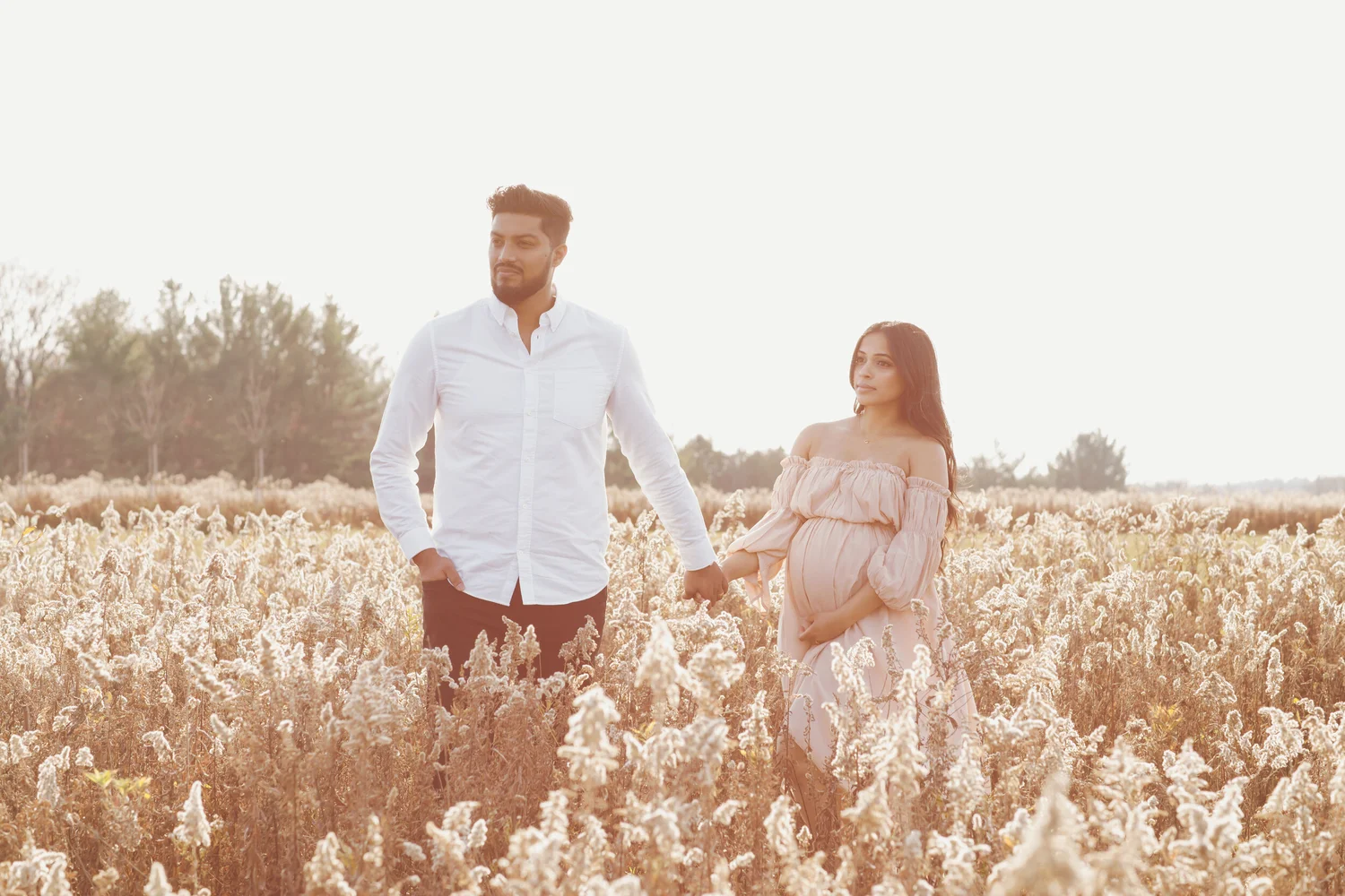An expectant couple walk hand in hand through a field of tall dried grasses, her hand on her bump.