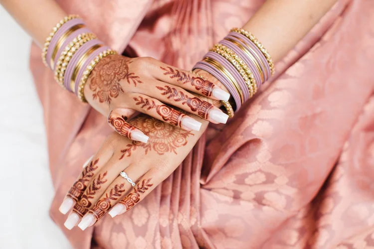 A bride's hennaed hands with stacked gold and lavender bangles and a diamond ring, resting on a blush-pink silk saree.