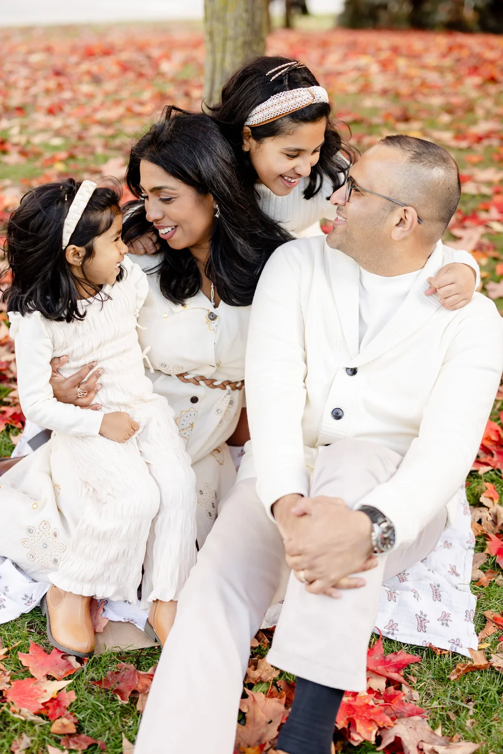 A family of four dressed in cream laugh together on the grass among red and orange autumn leaves.