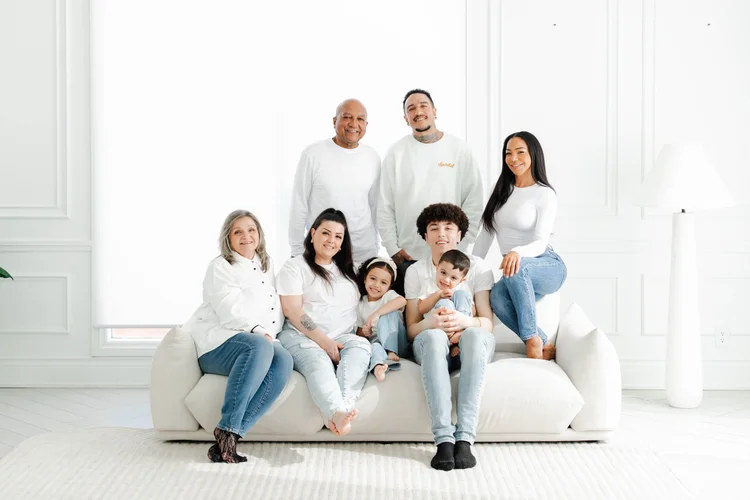 A large family dressed in white and denim, from grandparents to a toddler, gather on a white sofa in a bright, airy studio.