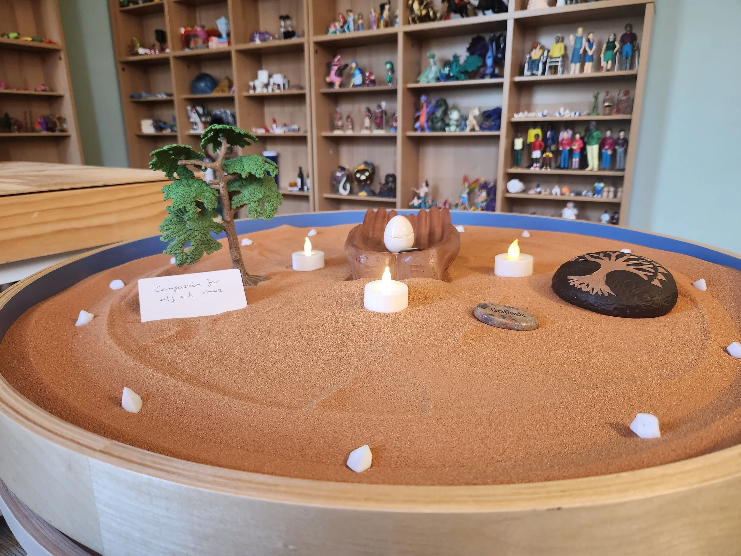 A round tray of warm sand holding a small tree, glowing candles, carved wooden hands cradling an egg, a stone engraved with a tree of life, and a handwritten card reading Compassion for self and others