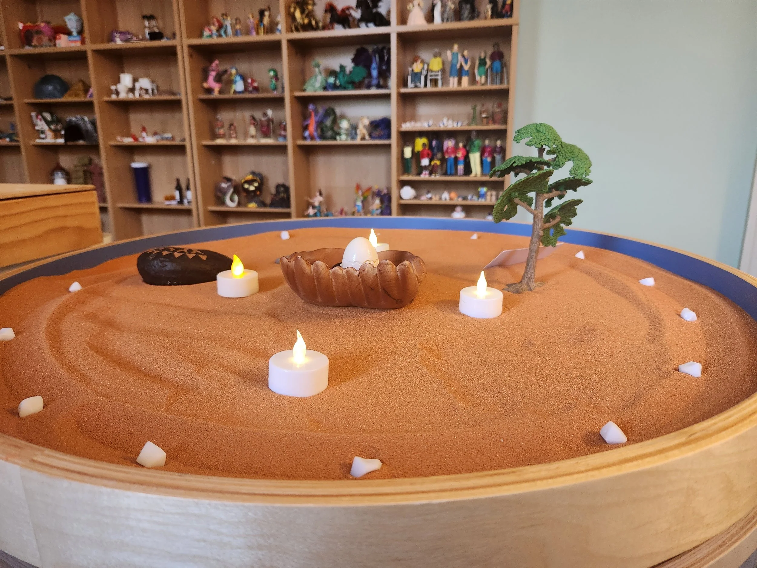 A wide view of a sand tray with candlelight and a small tree, with shelves of hundreds of miniature figures in the background