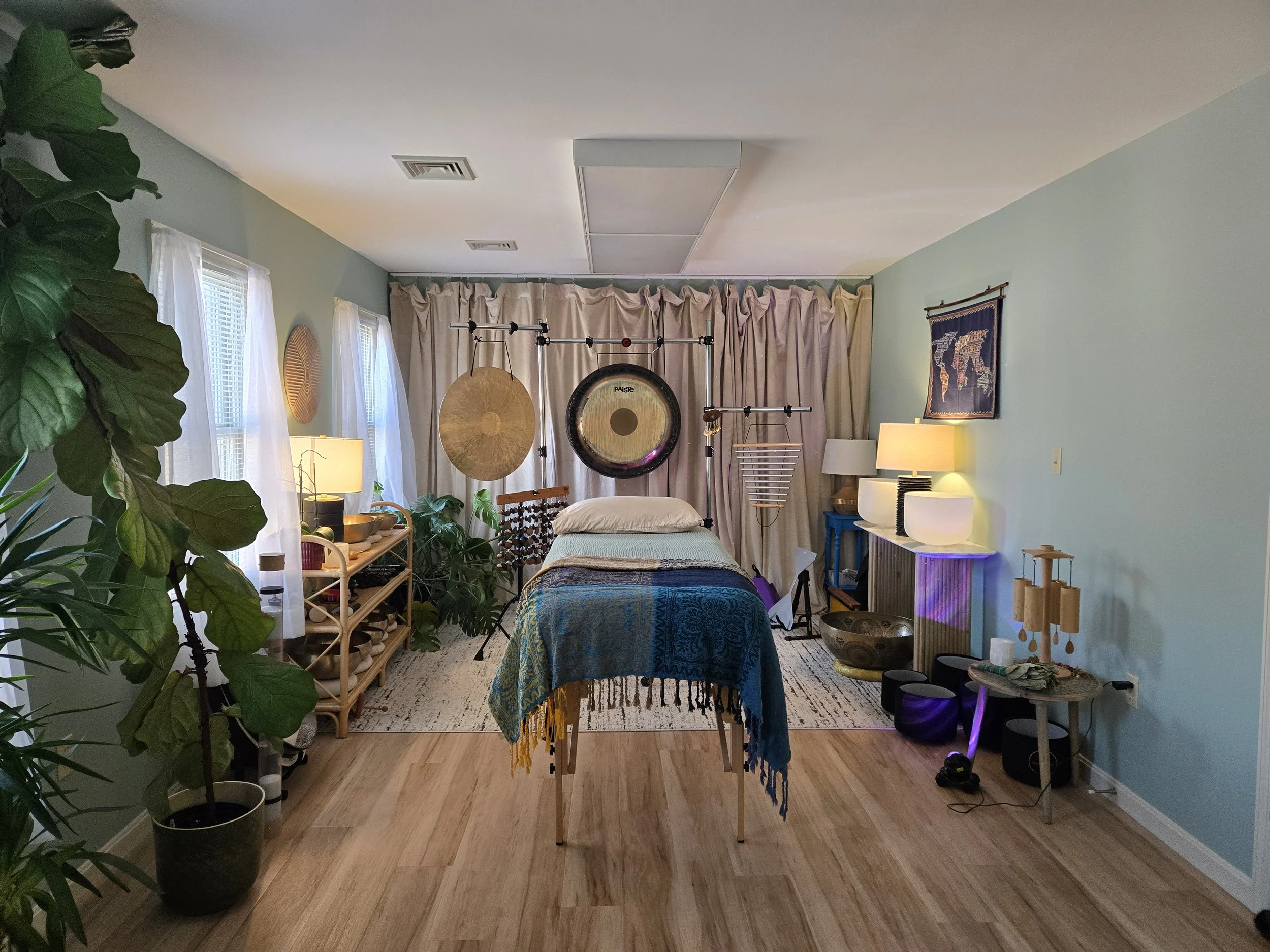 The sound and movement space at Meadow Grove Counseling, featuring gongs, crystal singing bowls, chimes, a treatment table with a teal blanket, large houseplants, and soft lighting