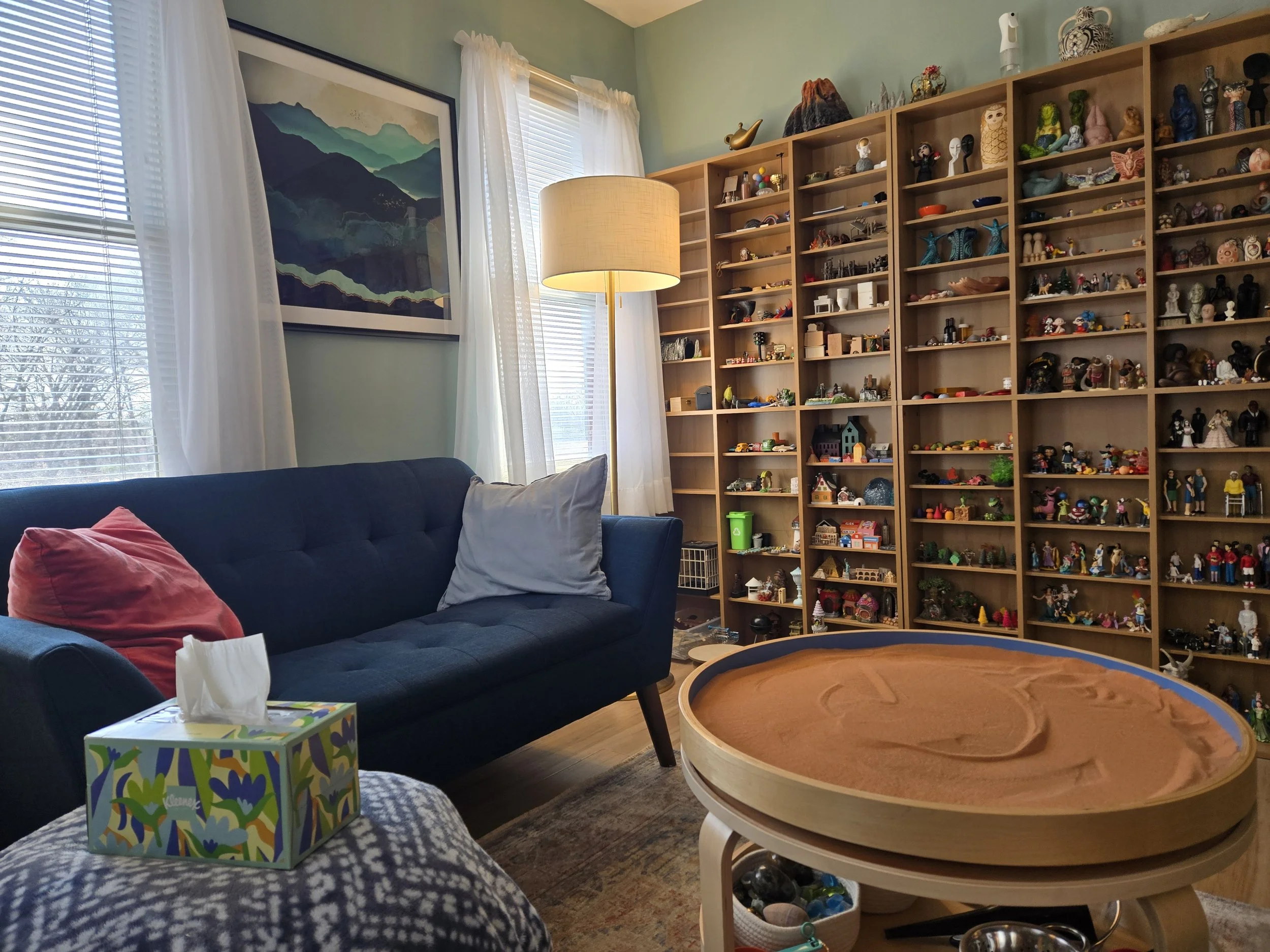 The therapy office at Meadow Grove Counseling in Lancaster PA. A sand tray sits in the foreground beside a blue sofa with warm pillows. Floor-to-ceiling shelves line the wall, filled with hundreds of sand tray miniatures. Natural light comes through sheer curtains. The space is warm, unhurried, and intentional.