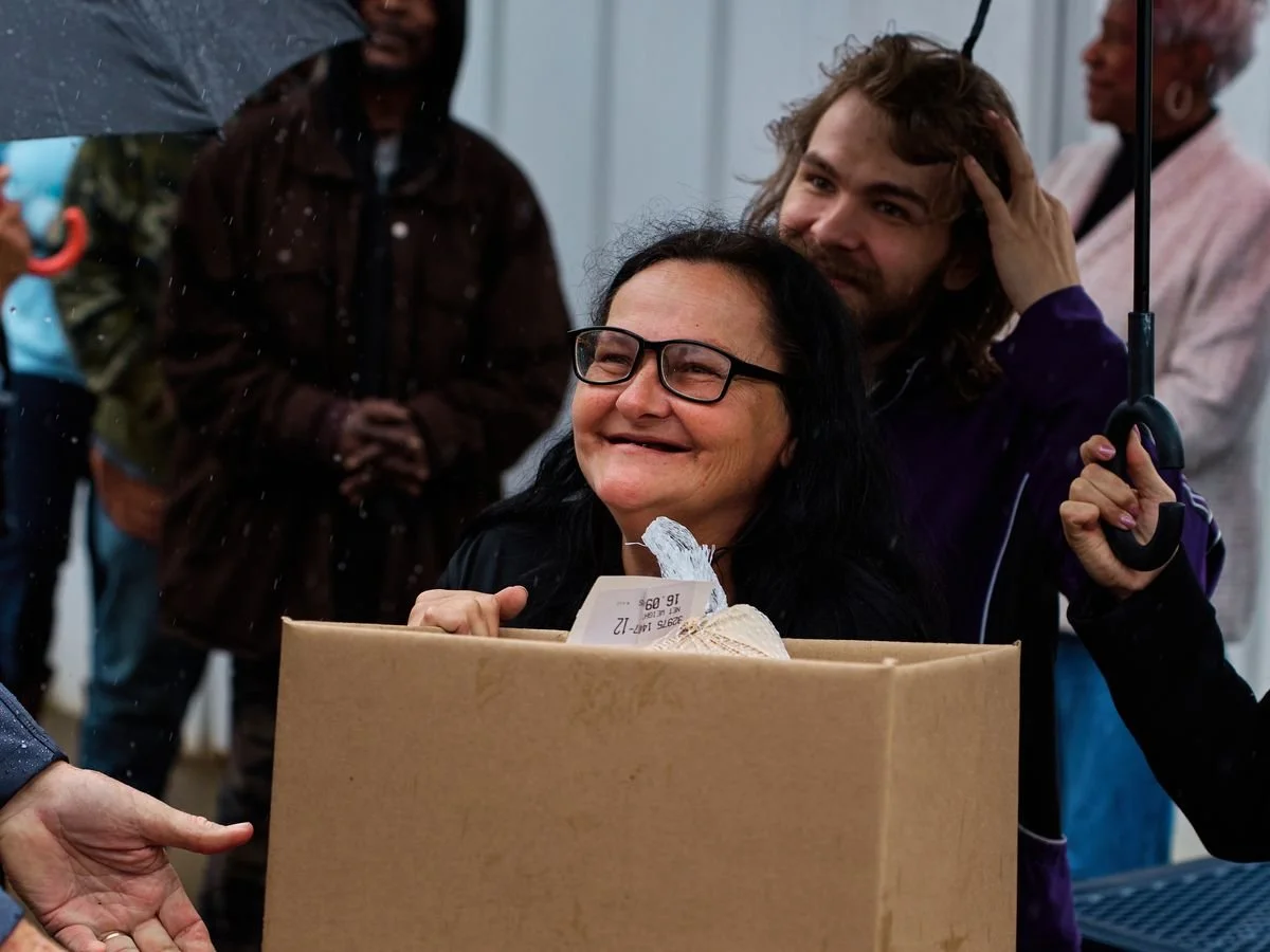 A neighbor smiles over her Gratitude Week meal box as volunteers gather behind her in the rain