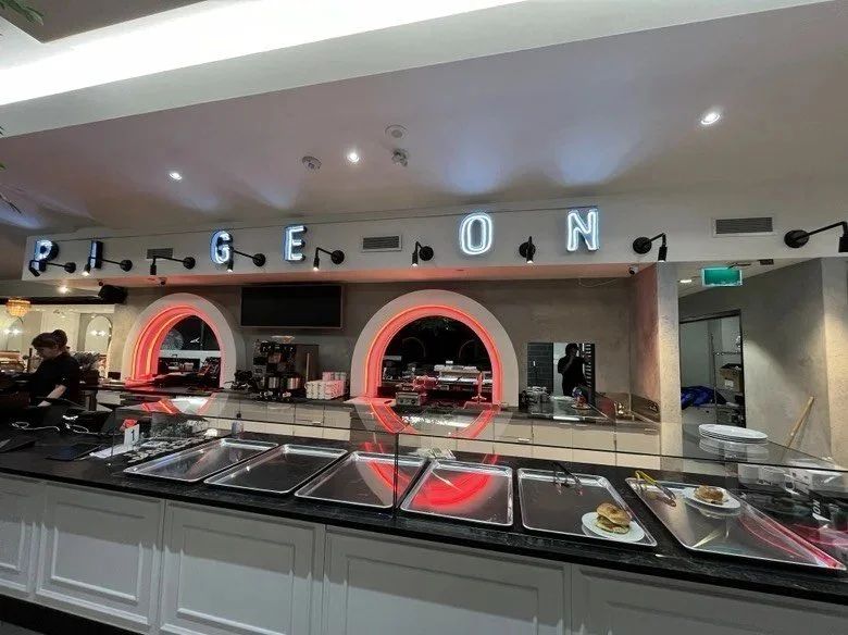 Self-serve café counter with glass food display, black stone countertop, illuminated 'PIGEON' signage, and two red-lit arched service windows behind the preparation area.