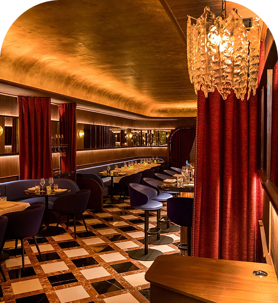 Upscale restaurant interior with warm lighting, curved seating booths, red velvet curtains, and a black-and-white checkered floor.