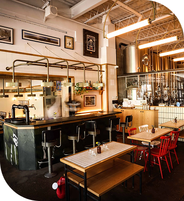 Industrial-style restaurant interior with an open bar, communal dining tables and exposed ceiling details.