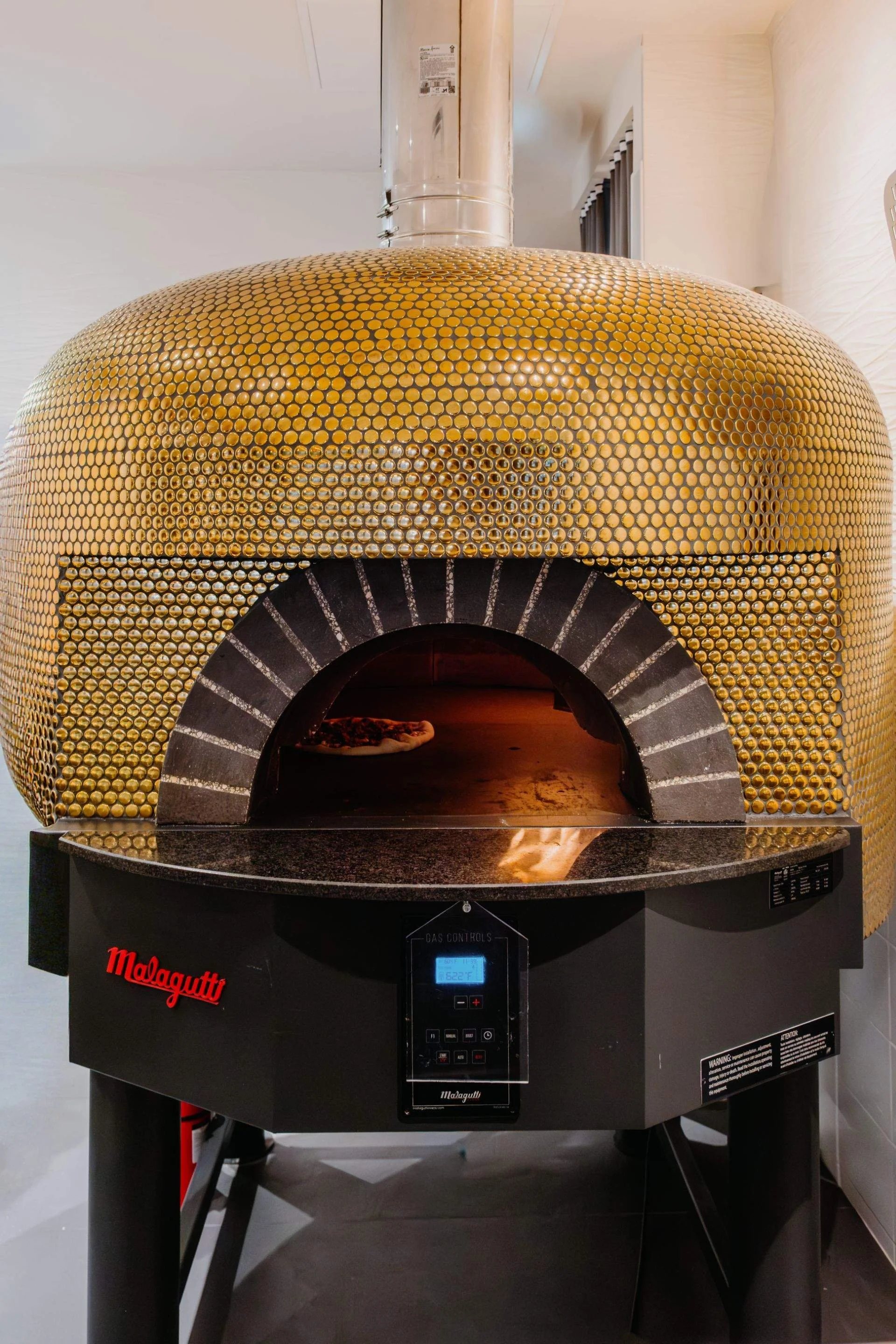 Large gold-tiled dome pizza oven with a stone-trimmed opening and pizza baking inside.