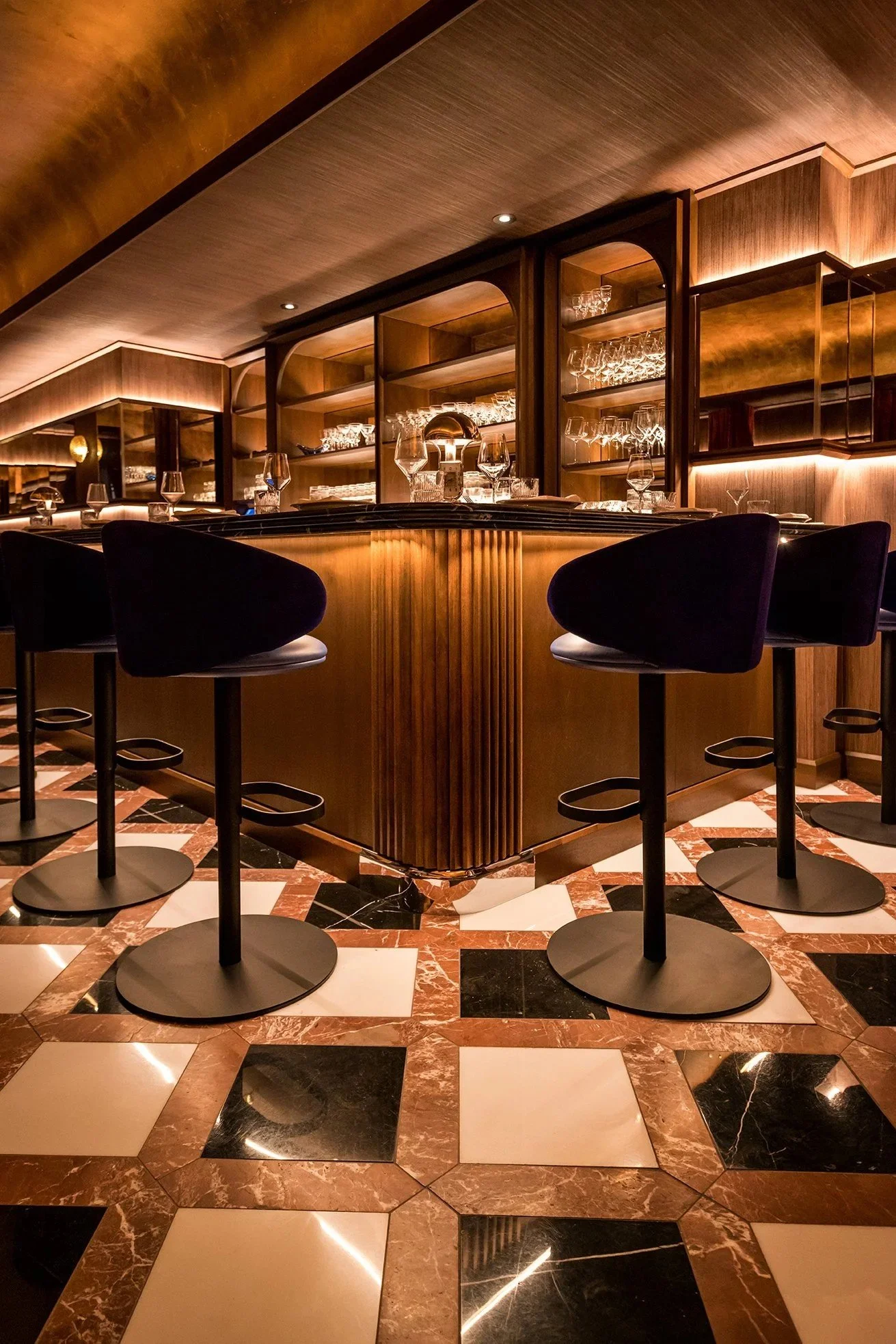 Wood-paneled bar with illuminated glass shelving, dark upholstered bar stools, and a black-and-white geometric marble tile floor in a warmly lit restaurant.