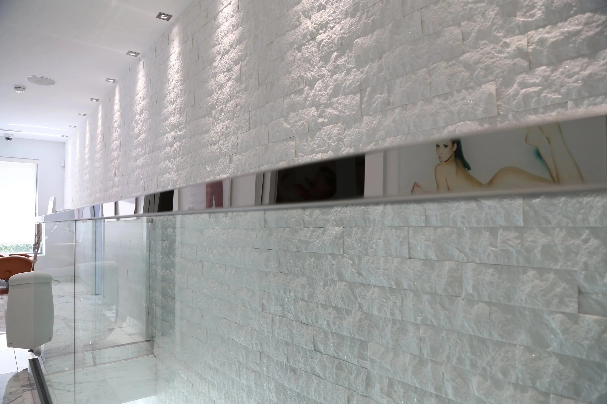 Bright upper-level hallway featuring textured white stone walls, glass railings, recessed lighting, and a clean, modern clinic design.