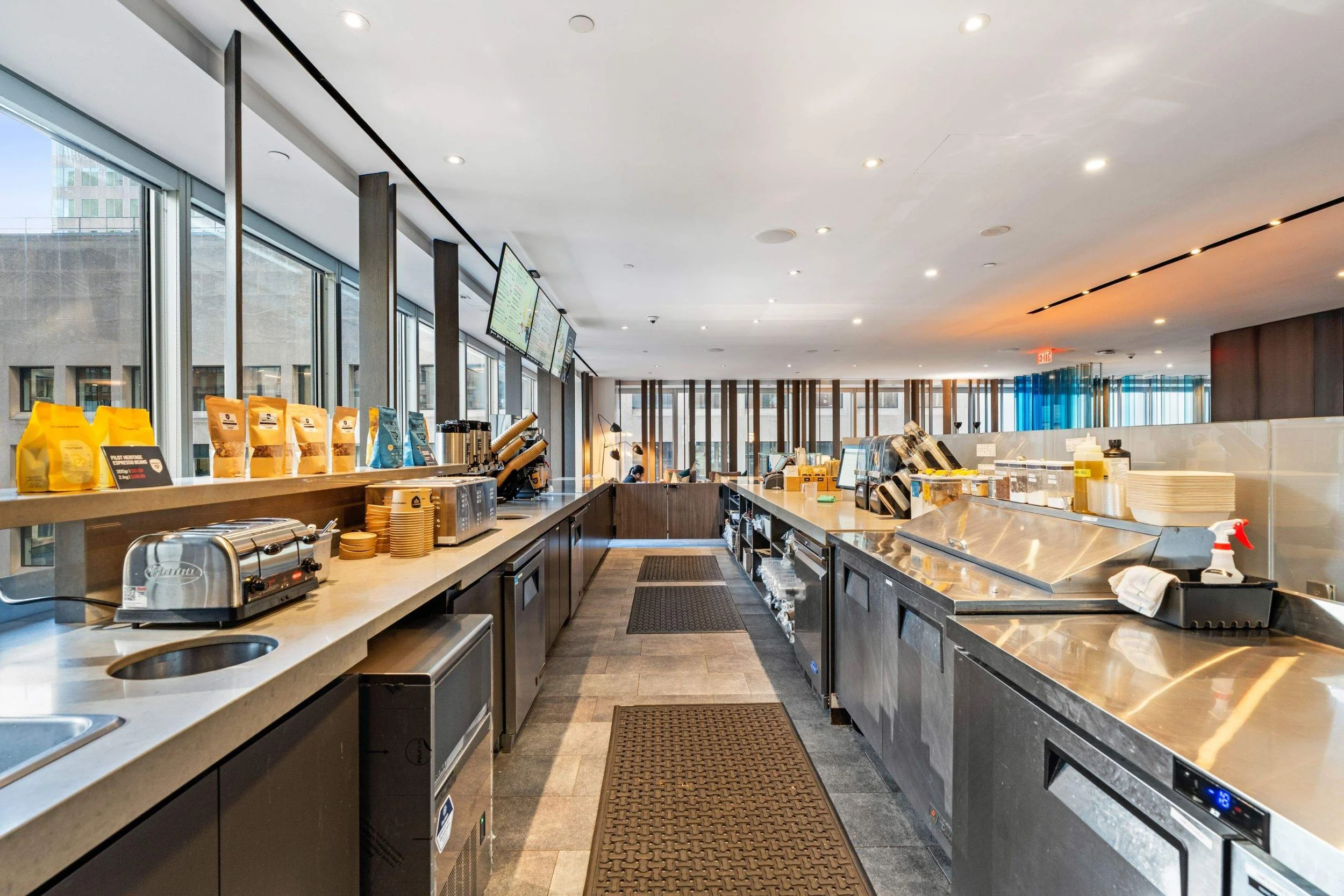 Behind-the-counter view of a modern café kitchen with stainless steel prep stations, equipment, and large windows.