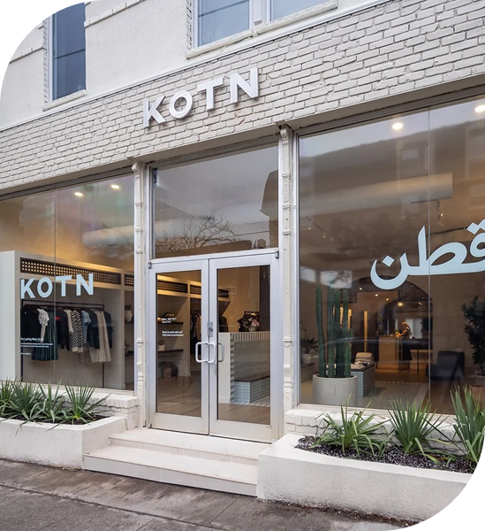 The exterior of a KOTN store location, with a brick wall, tall glass windows and door, and planters out front