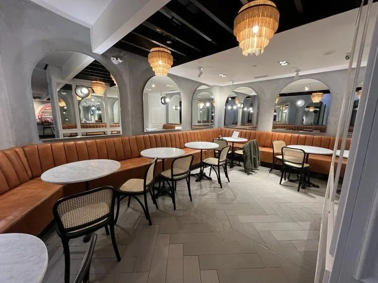 Bright café seating area with curved tan banquettes, marble café tables, arched mirrors, and decorative chandeliers in a modern, elegant interior.