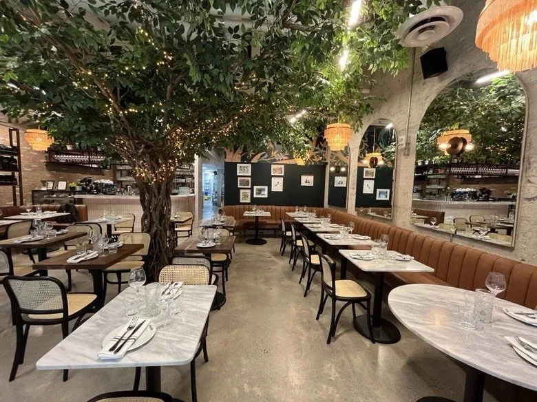 Bright restaurant dining area featuring marble-top tables, curved tan banquette seating, a large artificial tree with warm string lights, mirrored arches, chandeliers, and modern garden-inspired décor.