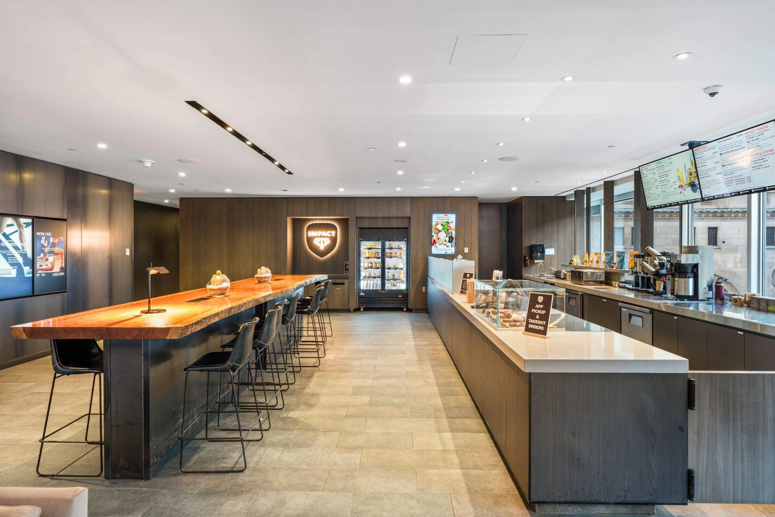 Wide view of a modern café with a long communal wood table, bar seating, service counter, and digital menu boards.