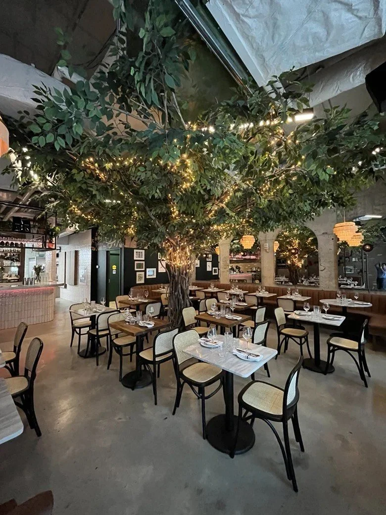 Restaurant dining area featuring a large artificial tree wrapped with warm string lights, marble-top tables, upholstered chairs, curved banquette seating, and mirrored archways creating an indoor garden atmosphere.
