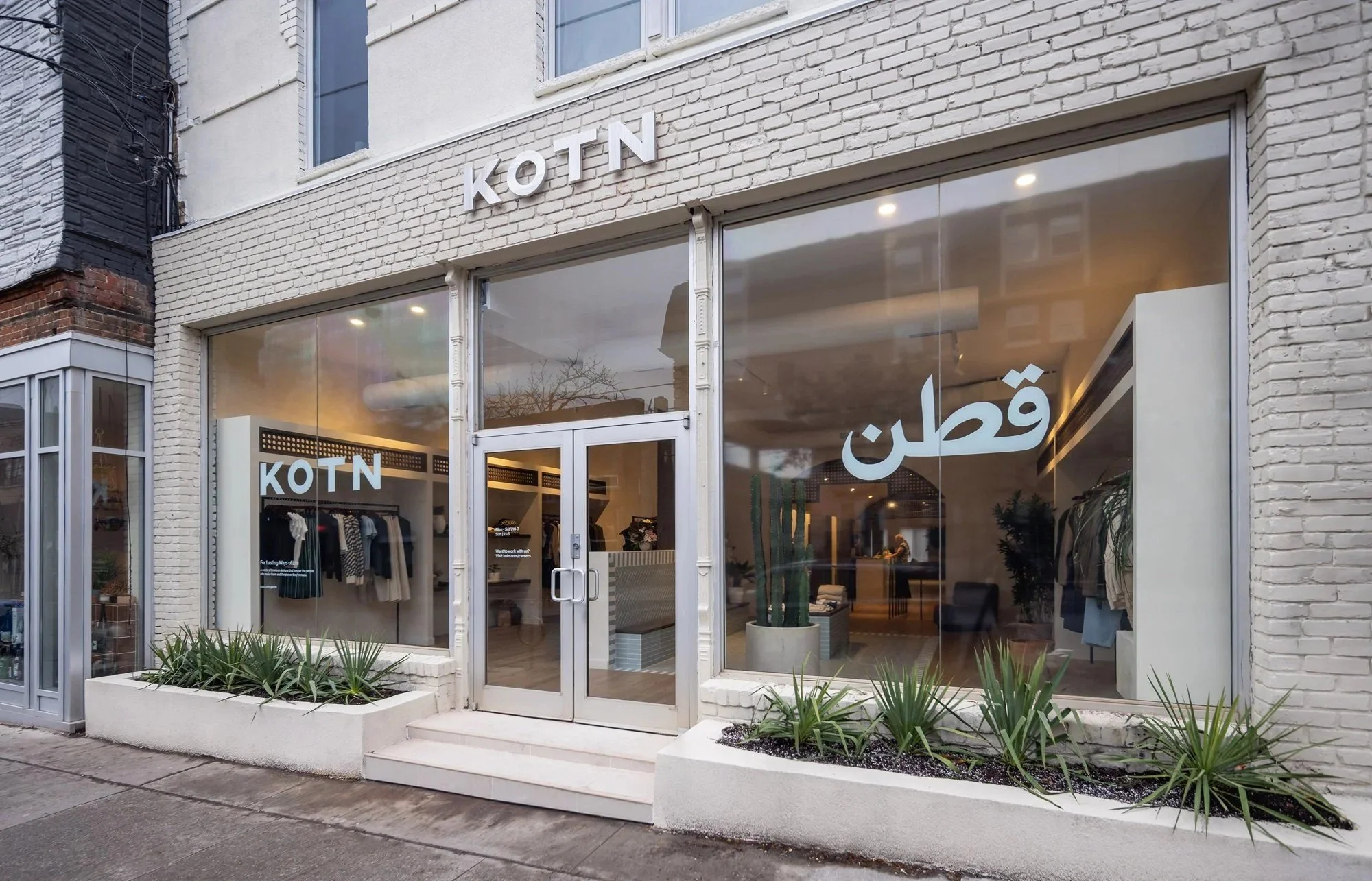 Street-level view of the KOTN storefront featuring large display windows, a light brick façade, minimalist signage, landscaped planters, and a warm, modern retail interior visible through the glass.