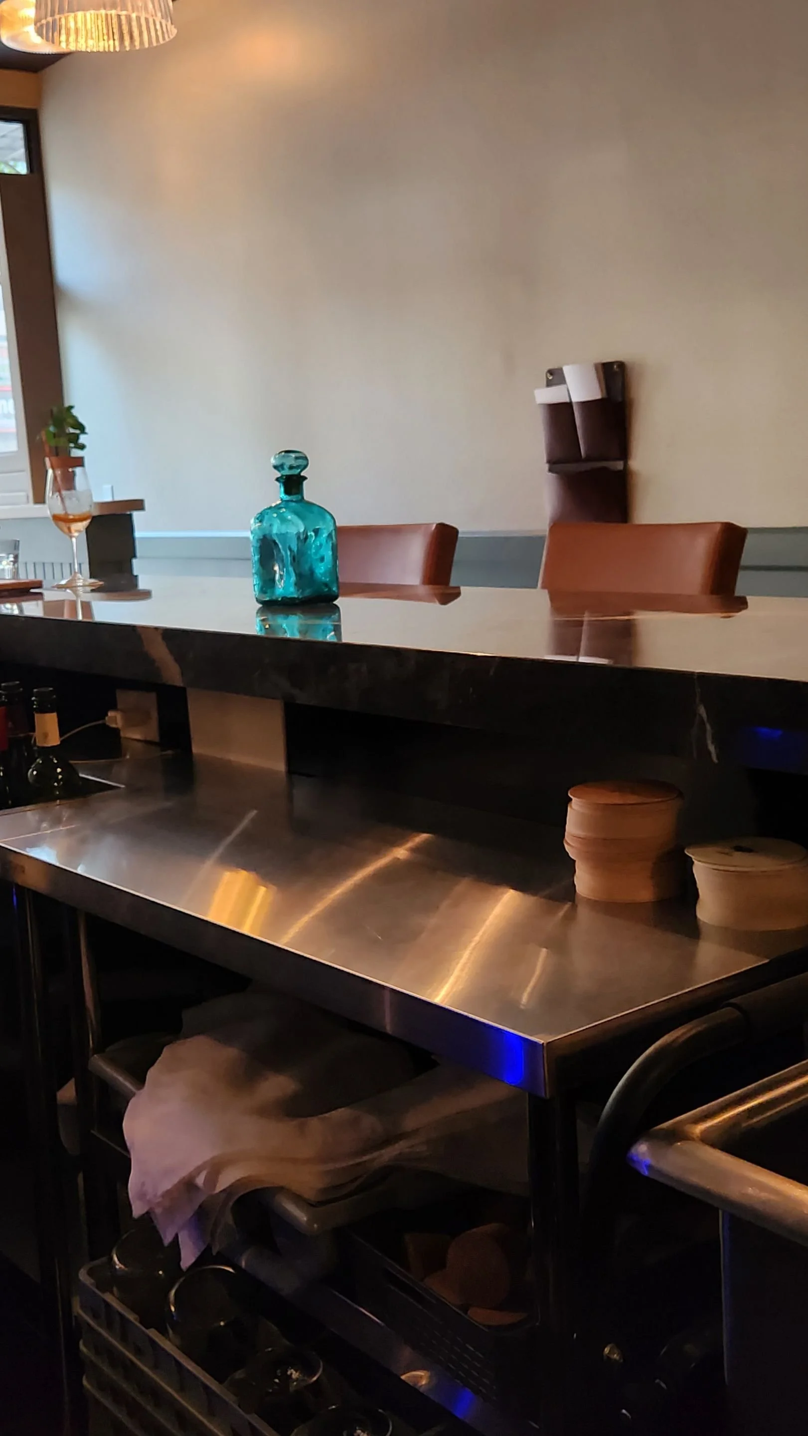 Restaurant bar with a polished black stone counter, leather stools, turquoise glass bottle, and stainless steel work area.