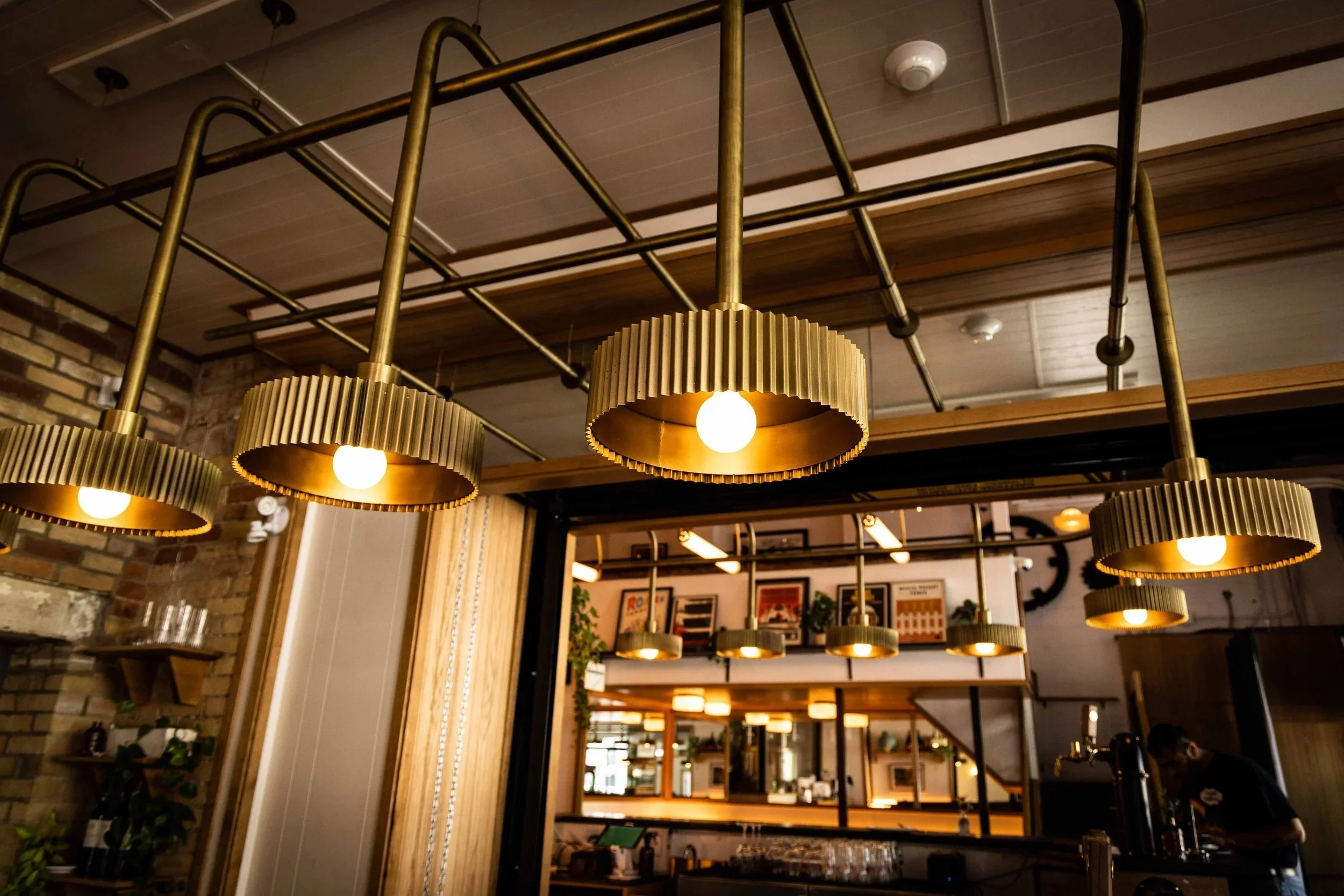 Close-up of brass pendant lights suspended above a restaurant bar, with warm ambient lighting, exposed brick, and reflections of the dining area in the background.