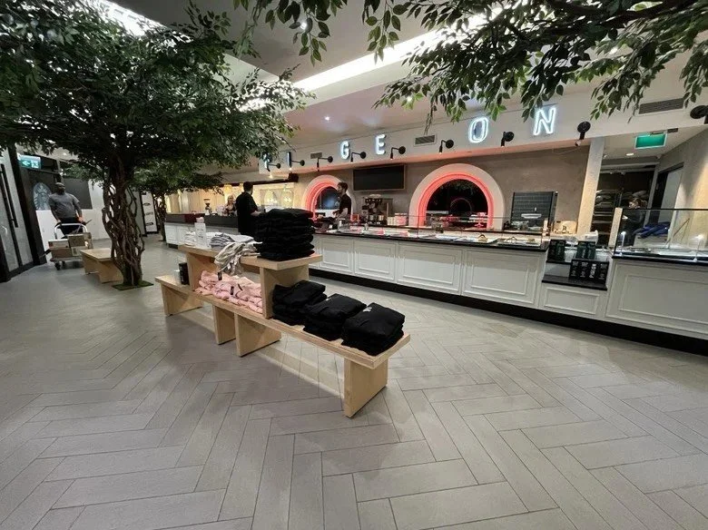Modern cafeteria serving area with illuminated food counters, arched display openings, indoor trees, and wooden benches displaying folded merchandise beneath overhead signage.
