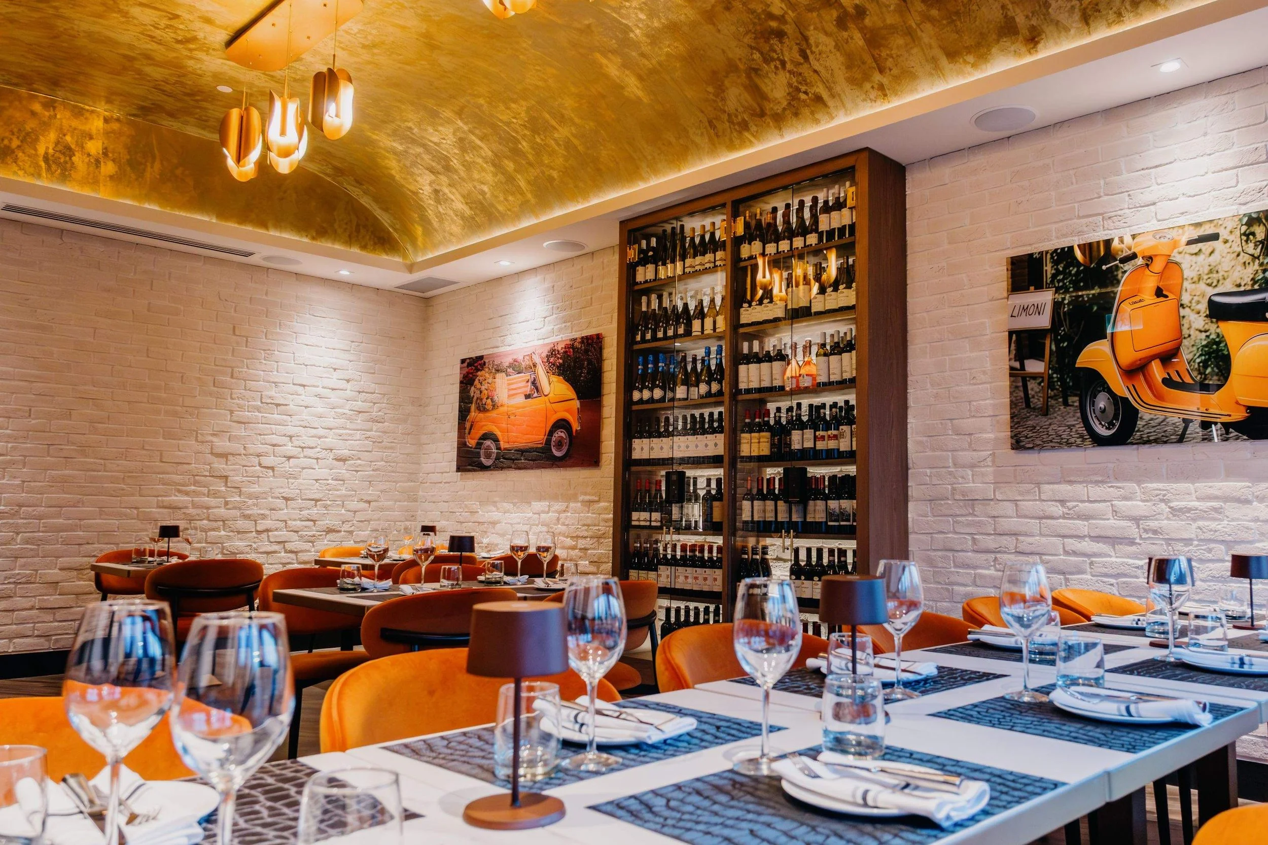 Warmly lit dining room with orange seating, white brick walls, a gold ceiling, and a tall glass-fronted wine display.