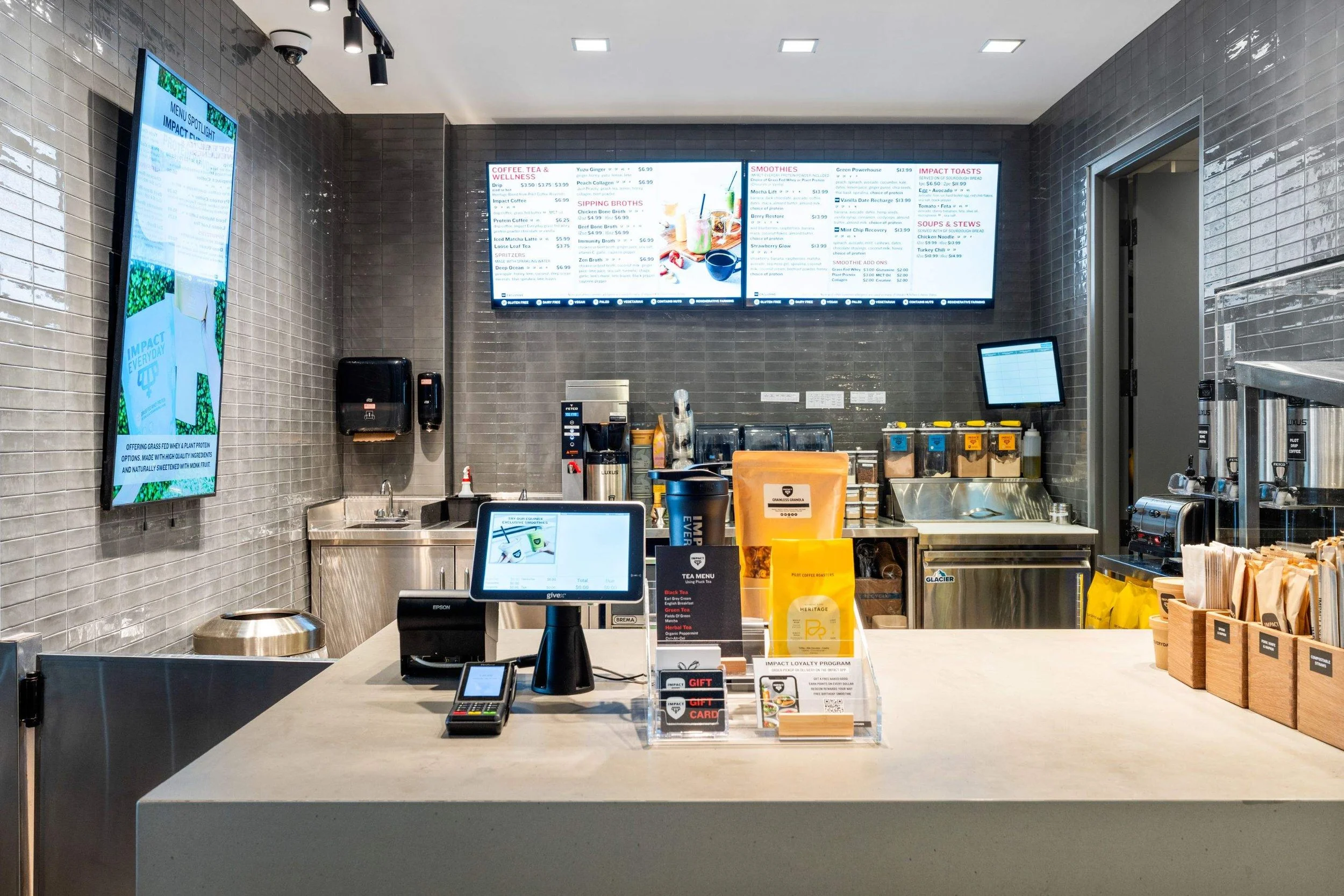 Impact Kitchen checkout counter with point-of-sale terminal, pastry display, digital menus, and café prep equipment.