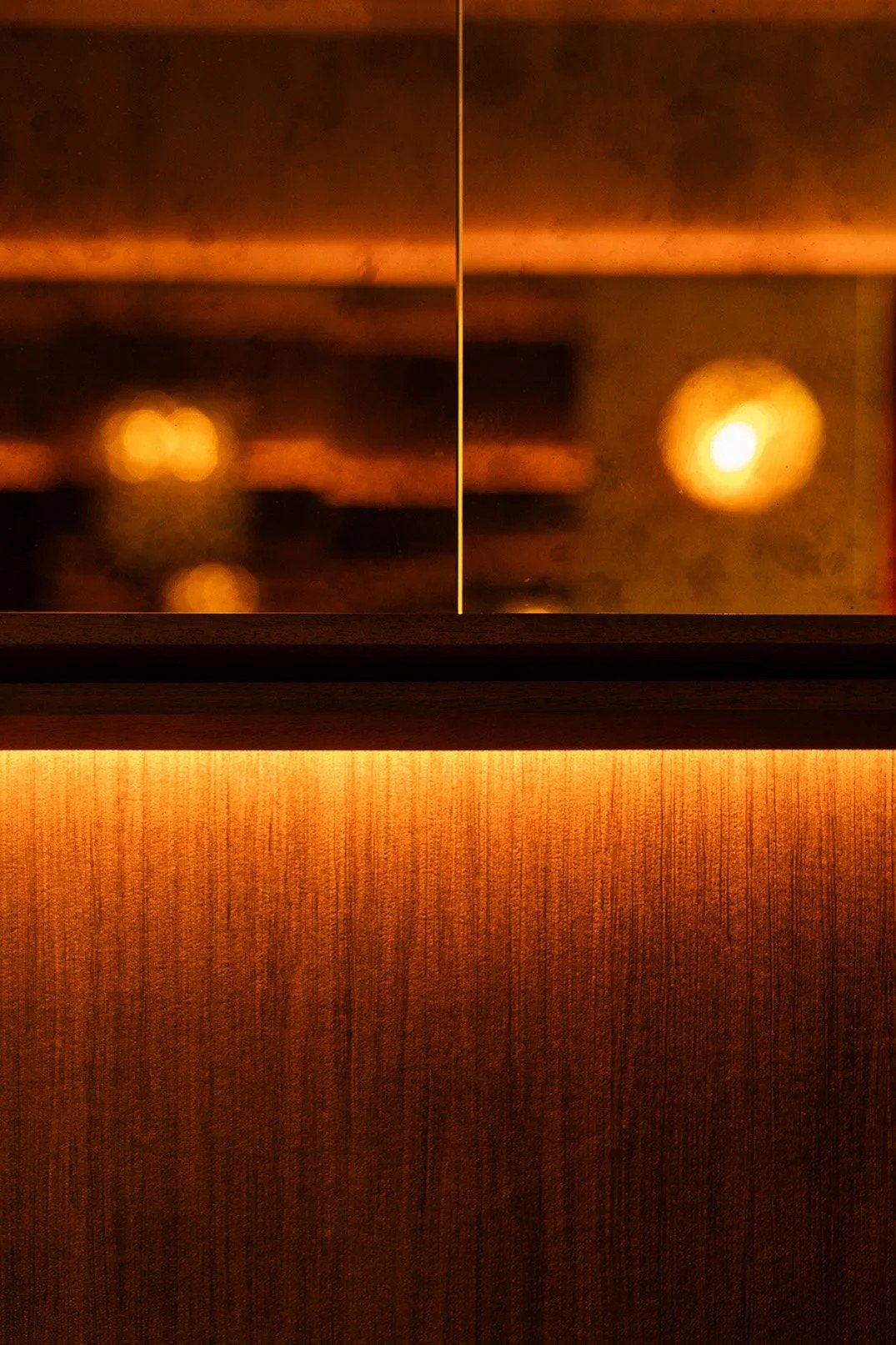Close-up of warm wood paneling beneath a mirrored wall with integrated accent lighting, reflecting softly blurred amber wall sconces in the background.