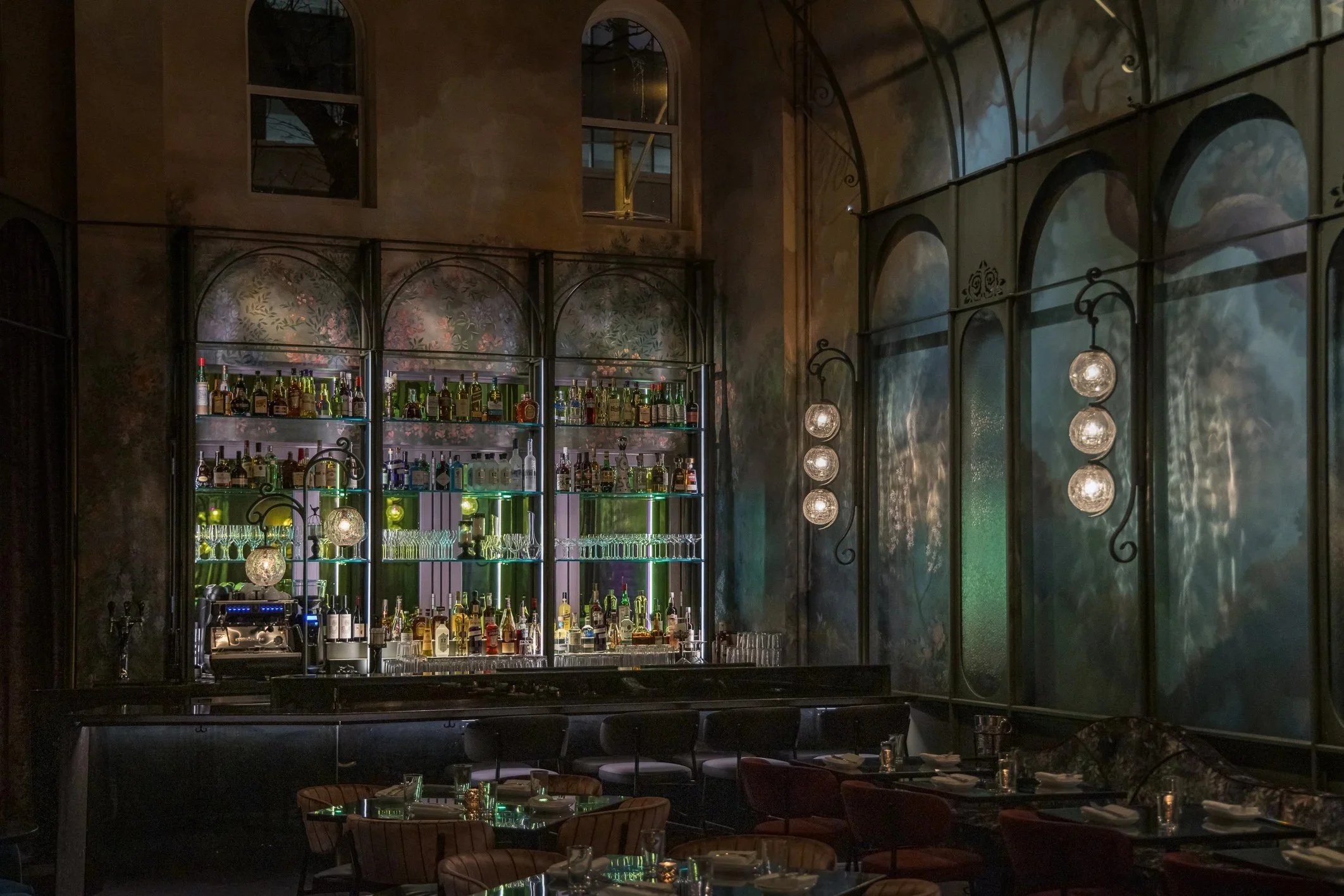Sophisticated bar and dining space with towering ceilings, backlit liquor shelves, painted botanical murals, arched architectural details, globe pendant lights, and intimate table seating.