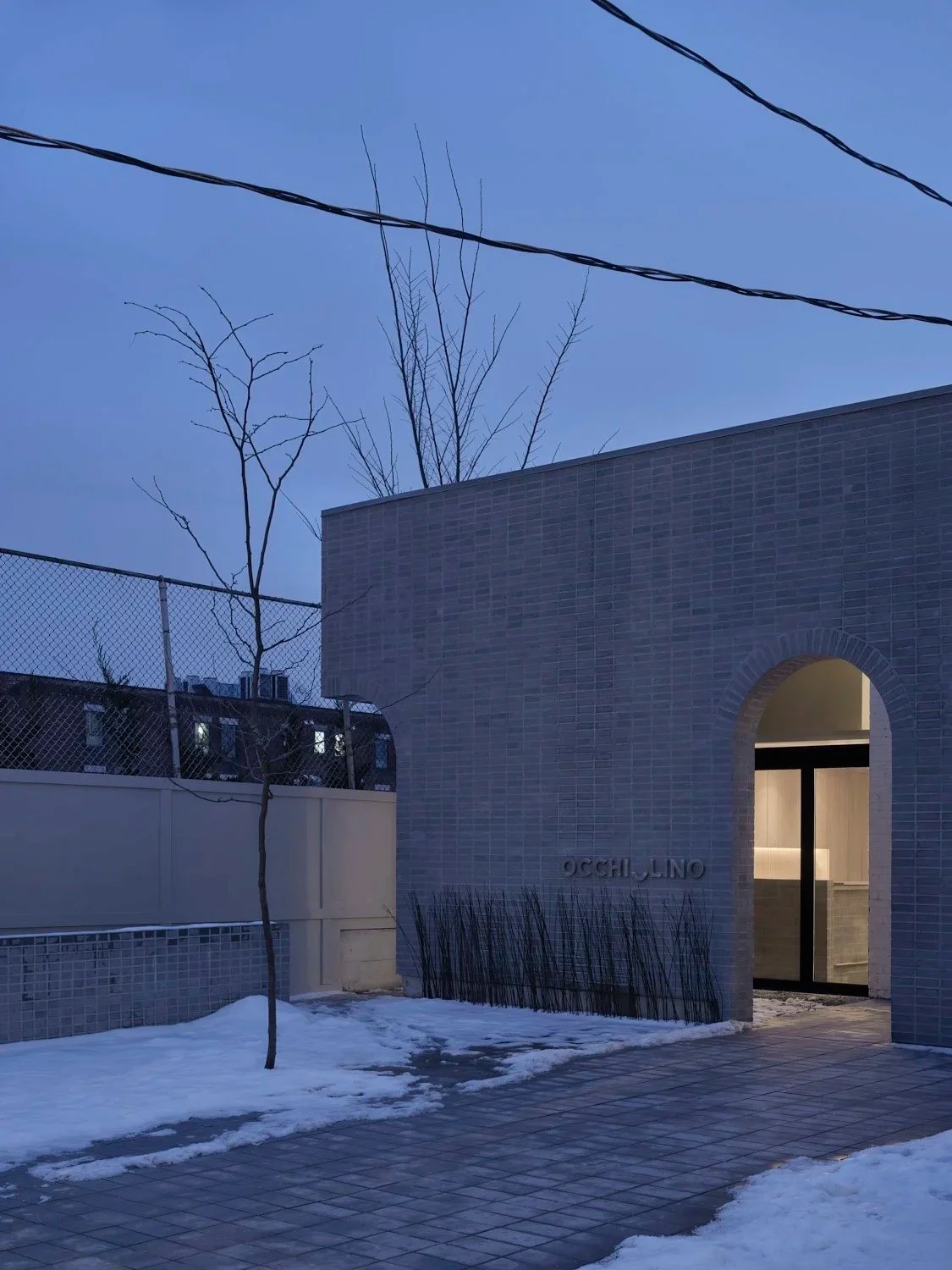 Exterior view of Occhiolino restaurant at dusk featuring a light grey brick facade, illuminated arched entrance, snow-covered courtyard, and minimalist landscaping.