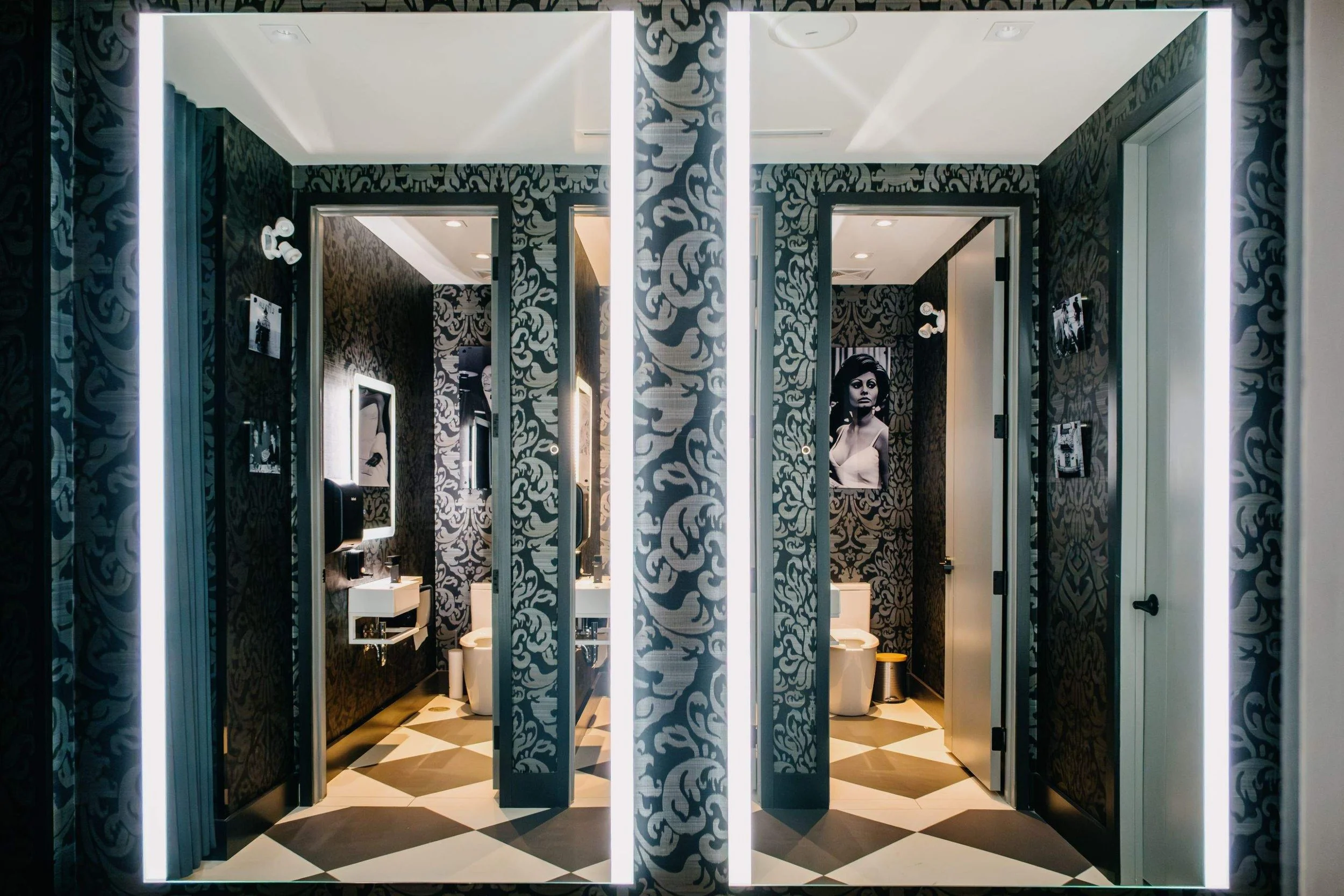 Two large illuminated mirrors reflecting restaurant restrooms decorated with dark patterned wallpaper and black-and-white photography.