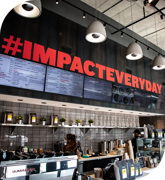 Modern café interior with large menu boards, hanging lights, and bold #IMPACTEVERYDAY signage above a service counter.