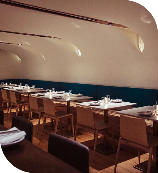 Minimalist restaurant dining area with curved white walls, built-in banquette seating, and neatly set wooden tables.