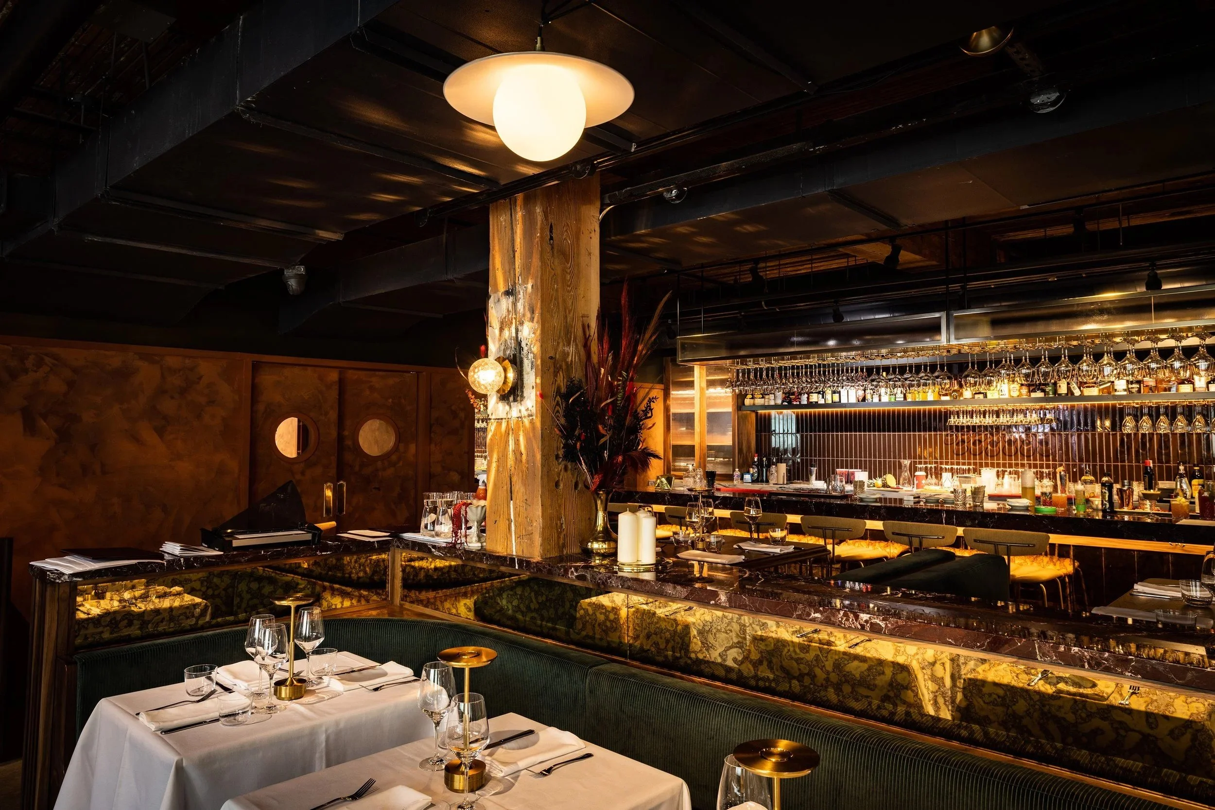 Upscale restaurant interior featuring elegant table settings, green banquette seating, a warmly lit marble bar, and exposed wood and industrial architectural details.