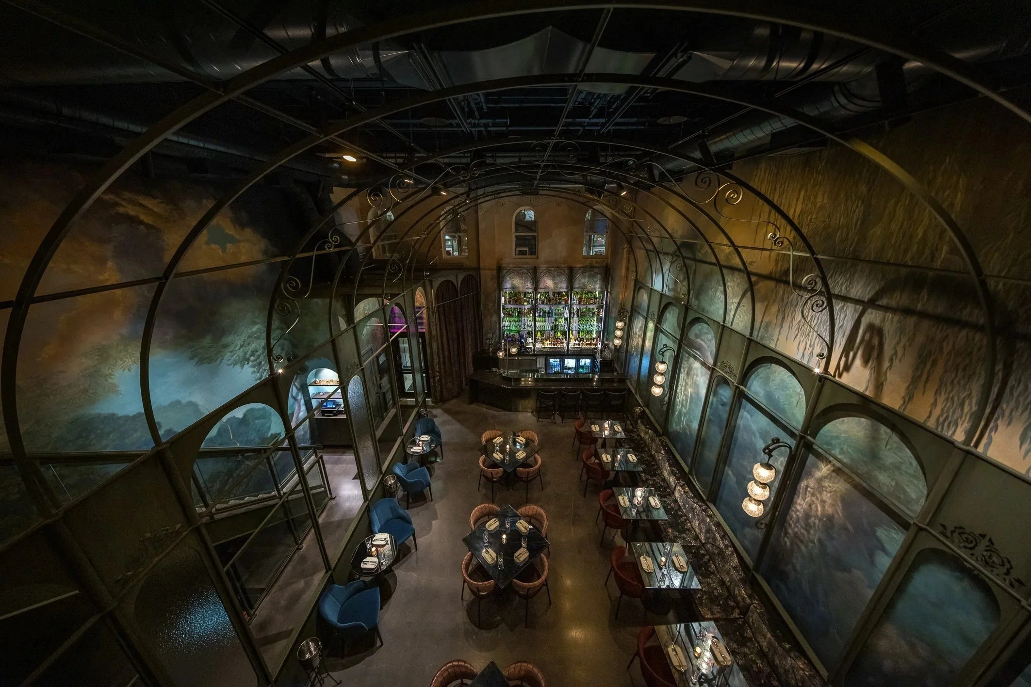Overhead view of a grand two-story restaurant with arched metal framework, dramatic hand-painted landscape murals, elegant dining tables, jewel-toned seating, and a backlit bar anchoring the space.