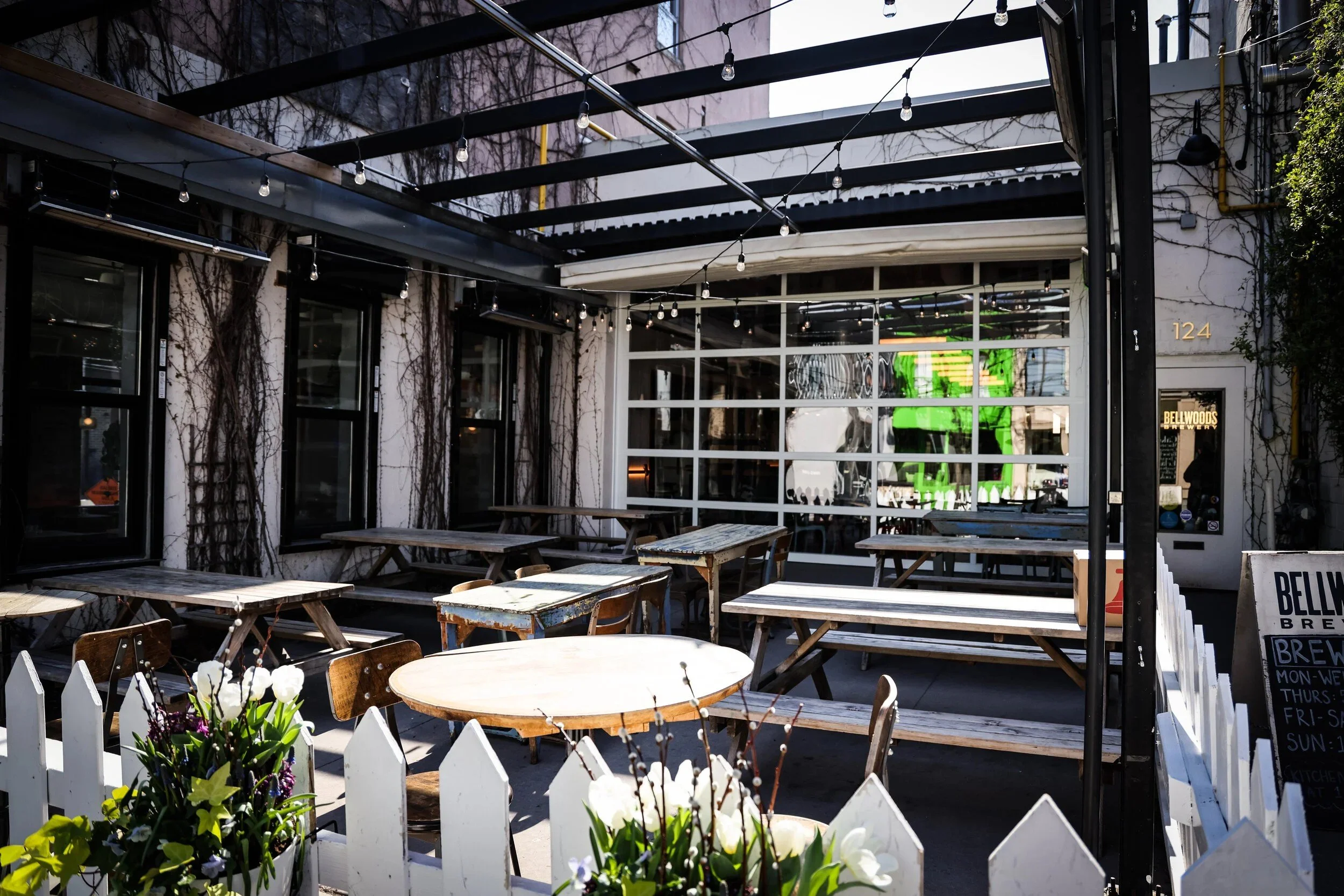 Outdoor brewery patio with wooden picnic tables beneath a black steel pergola strung with café lights, surrounded by climbing vines, large windows, and a white picket fence with flowering planters.