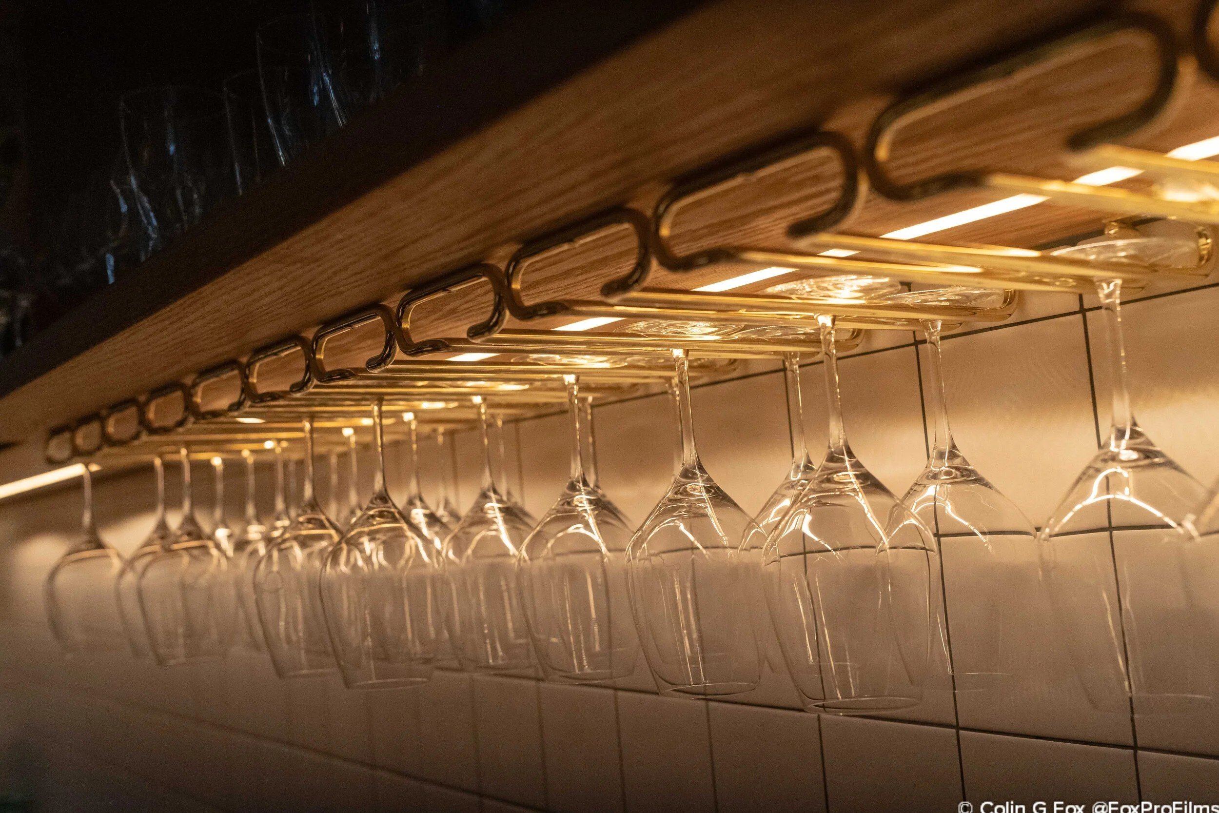 Rows of hanging wine glasses stored beneath a wooden shelf with warm under-cabinet lighting behind a restaurant bar.