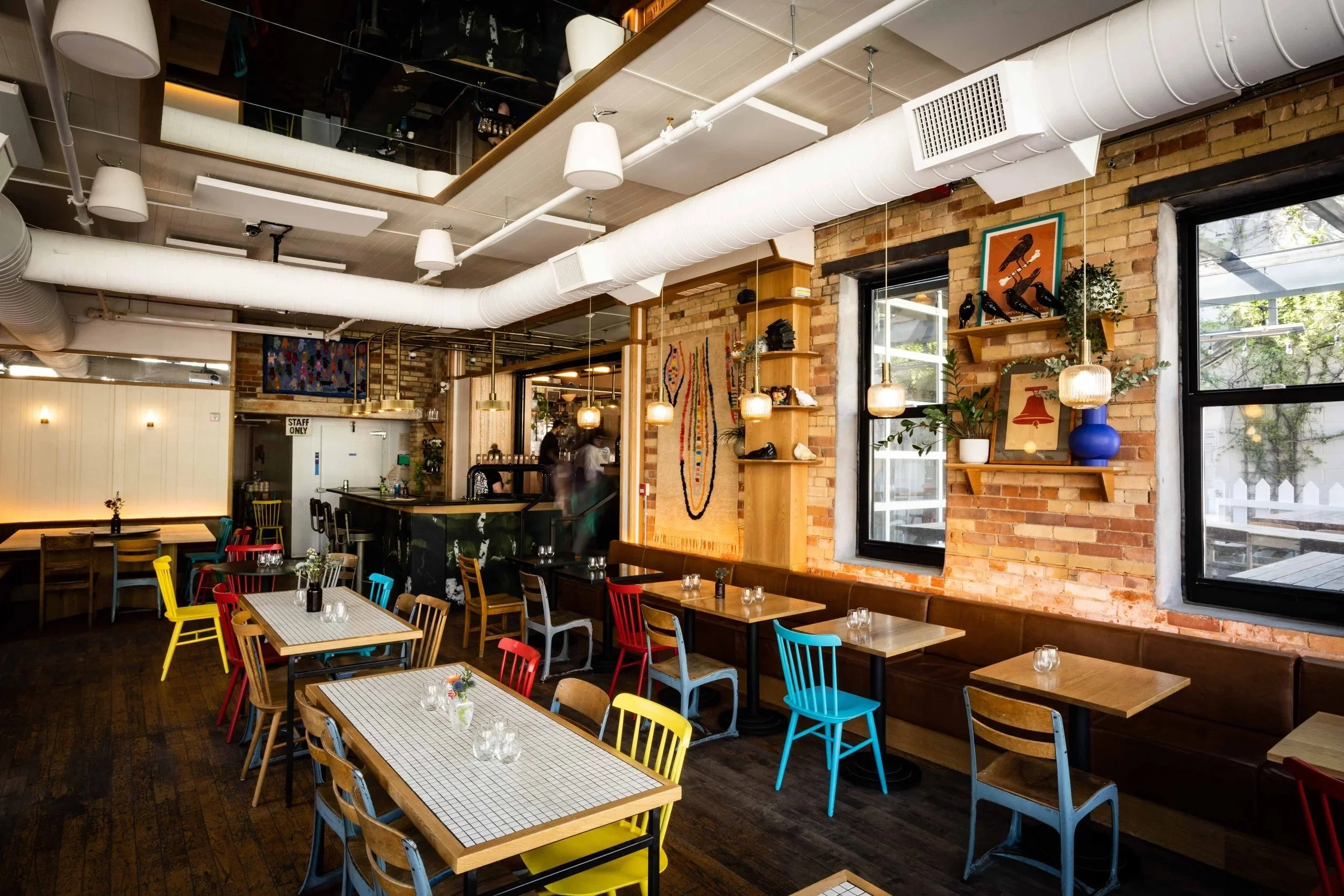 Restaurant dining area with exposed brick walls, colorful chairs, booth seating, pendant lights, and wood shelving with decorative accents.