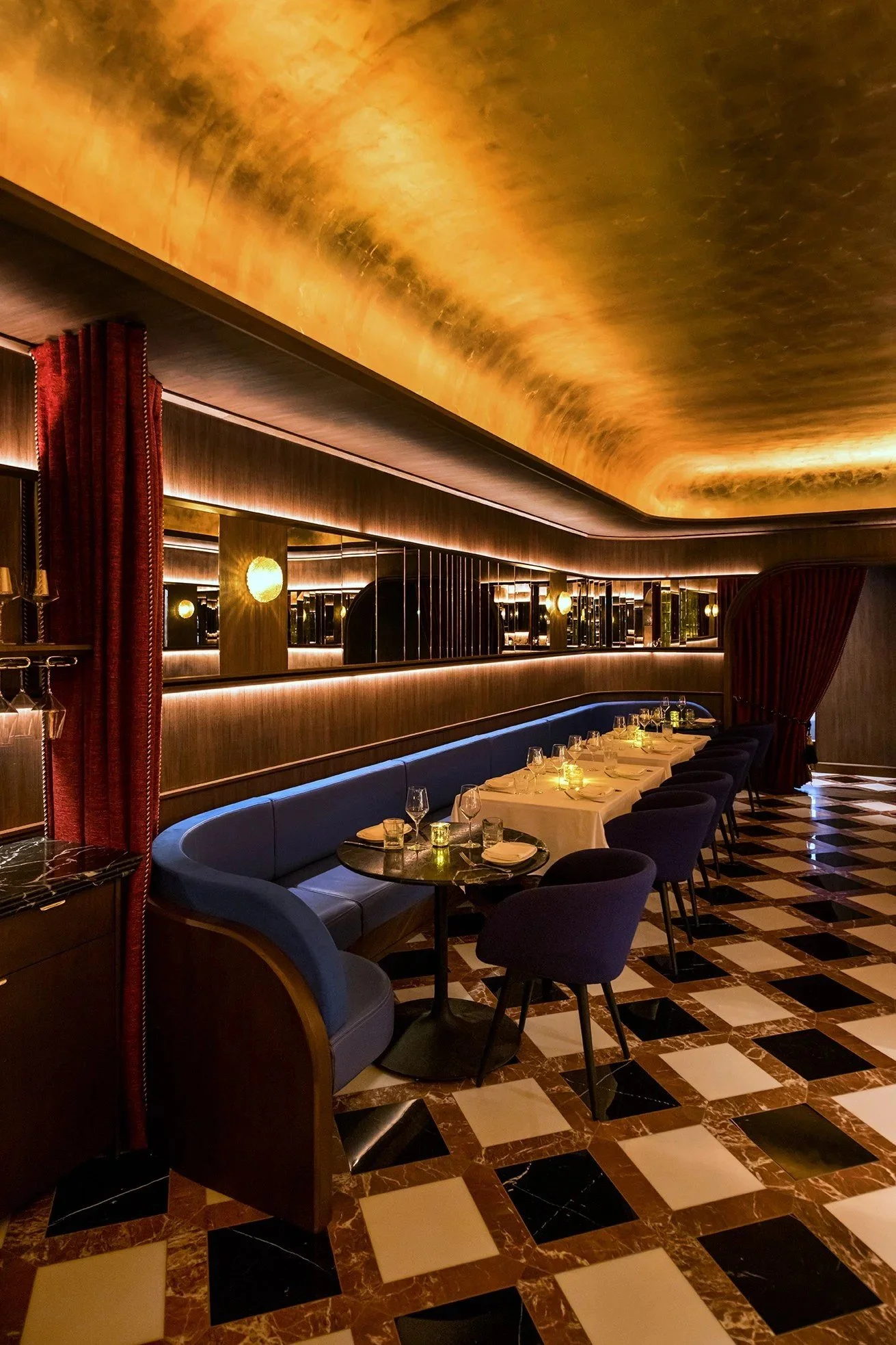 Sophisticated restaurant interior with curved blue banquette seating, white tablecloth dining tables, mirrored wood-paneled walls, a glowing gold-toned ceiling, and a black-and-white geometric tile floor.