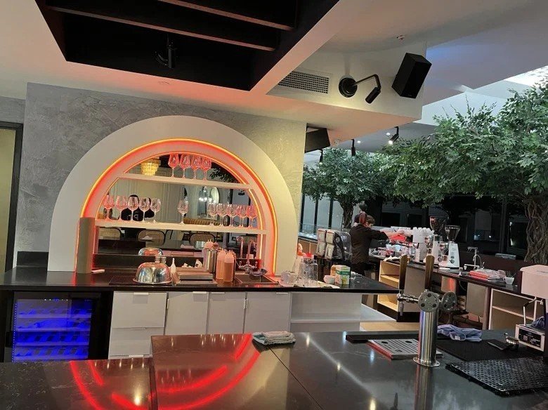 Contemporary café bar with a glowing arched back bar, black stone countertop, espresso equipment, and indoor trees creating a bright, modern hospitality space.