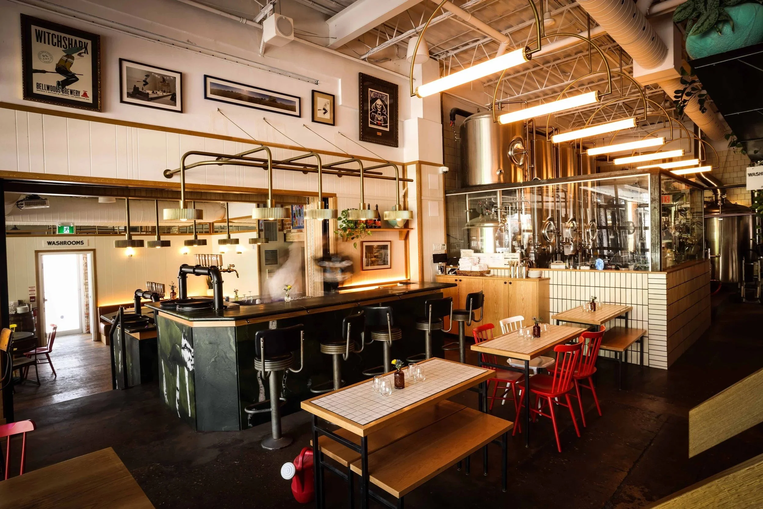 Wide view of a modern brewery and restaurant featuring a central bar with industrial stools, tiled dining tables, exposed brewing tanks behind glass, suspended brass lighting, and warm wood and metal finishes throughout.