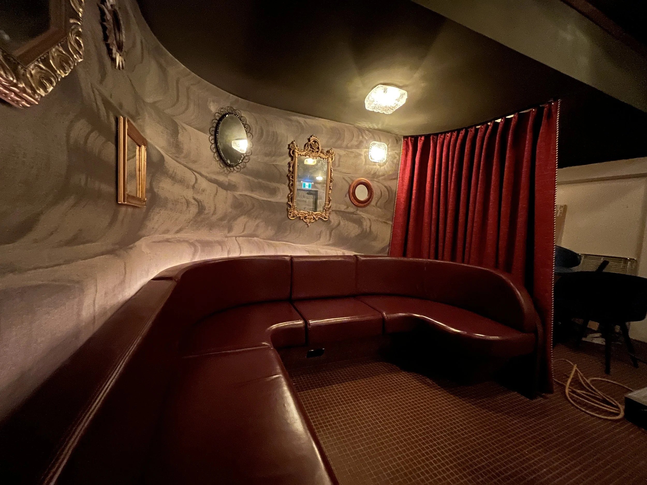Curved burgundy leather banquette wraps around a private dining nook with textured walls, vintage-style mirrors, warm sconces, and a red velvet curtain.