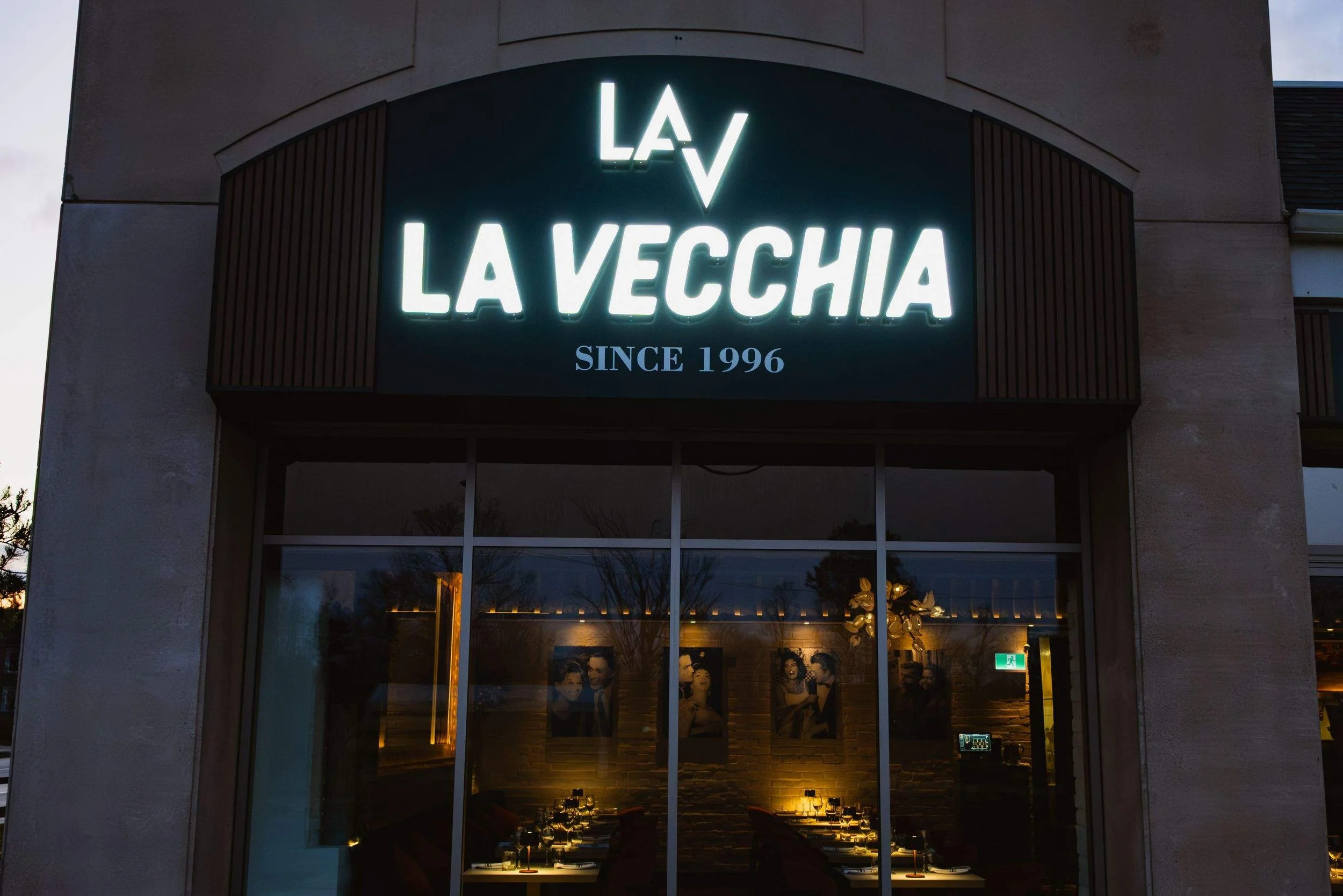 Exterior of La Vecchia restaurant with a large illuminated “La Vecchia Since 1996” sign above the front windows.