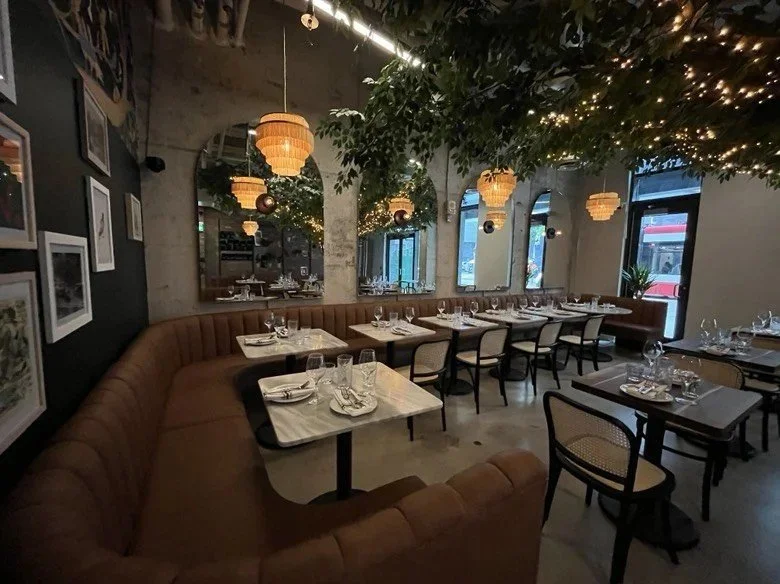 Restaurant dining area with curved tan banquette seating, marble-top tables, woven pendant chandeliers, mirrored arches, a large artificial tree with warm string lights, and floor-to-ceiling windows along the perimeter.