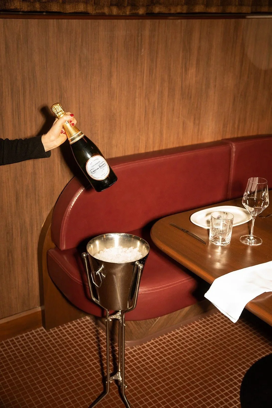 A hand holding a bottle of champagne beside a red leather banquette, with a chilled ice bucket on a stand and a restaurant table set with plates, wine glasses, and folded napkins against warm wood paneling.