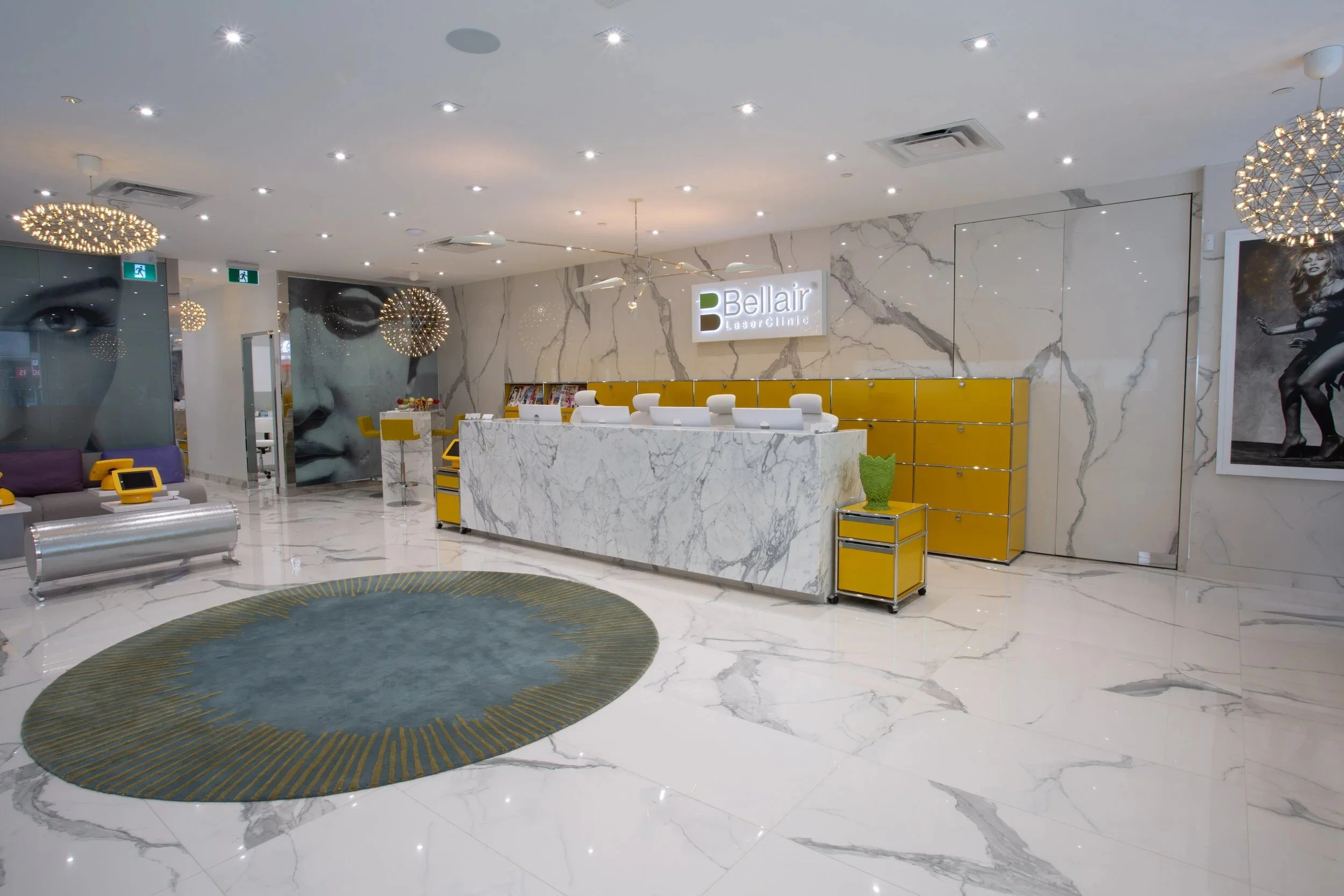 Modern Bellair Laser Clinic reception area with marble finishes, illuminated logo, white reception desk, yellow cabinetry, and contemporary waiting lounge.