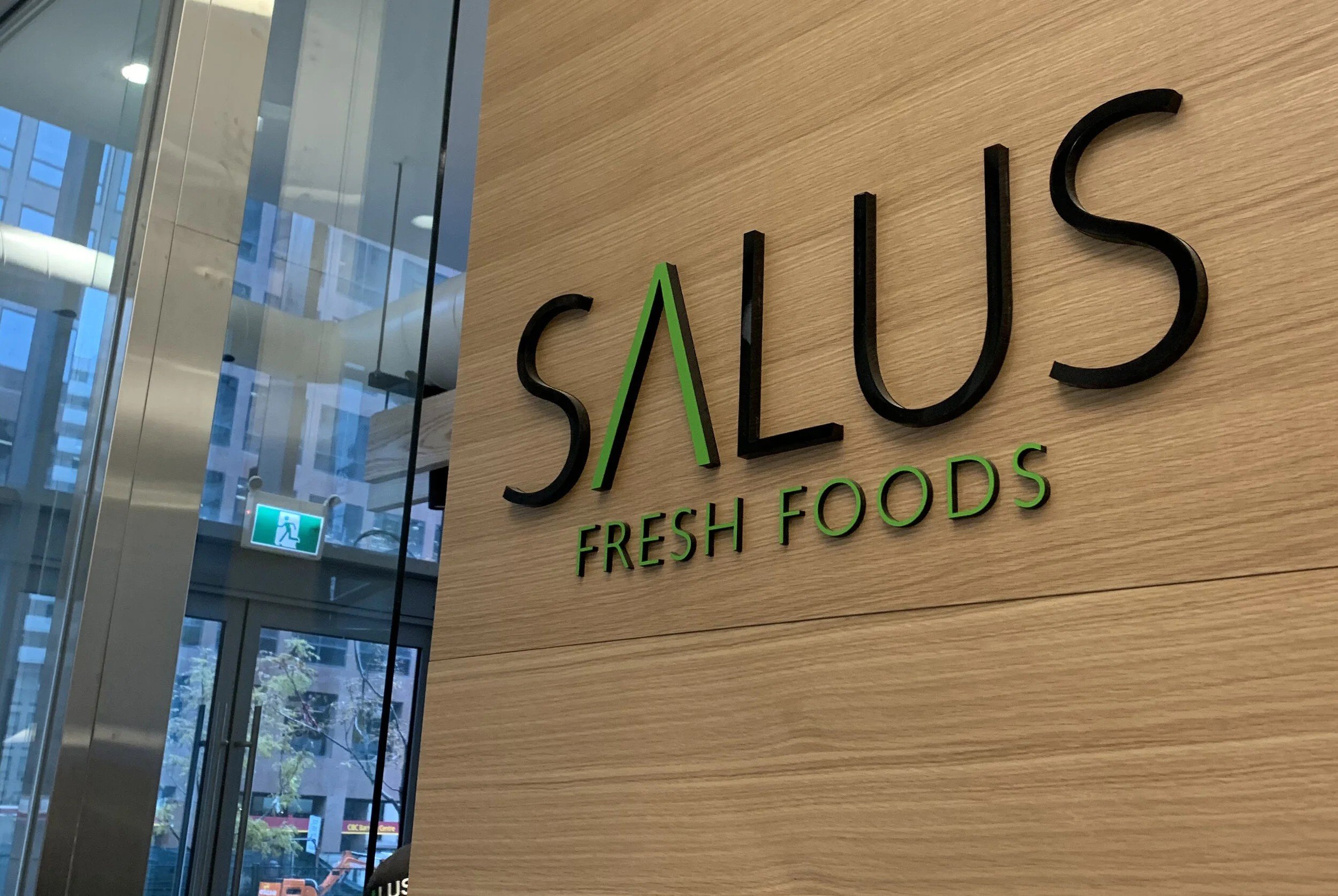 Salus Fresh Foods wall logo mounted on a light wood feature wall beside a glass storefront entrance in a modern commercial interior.